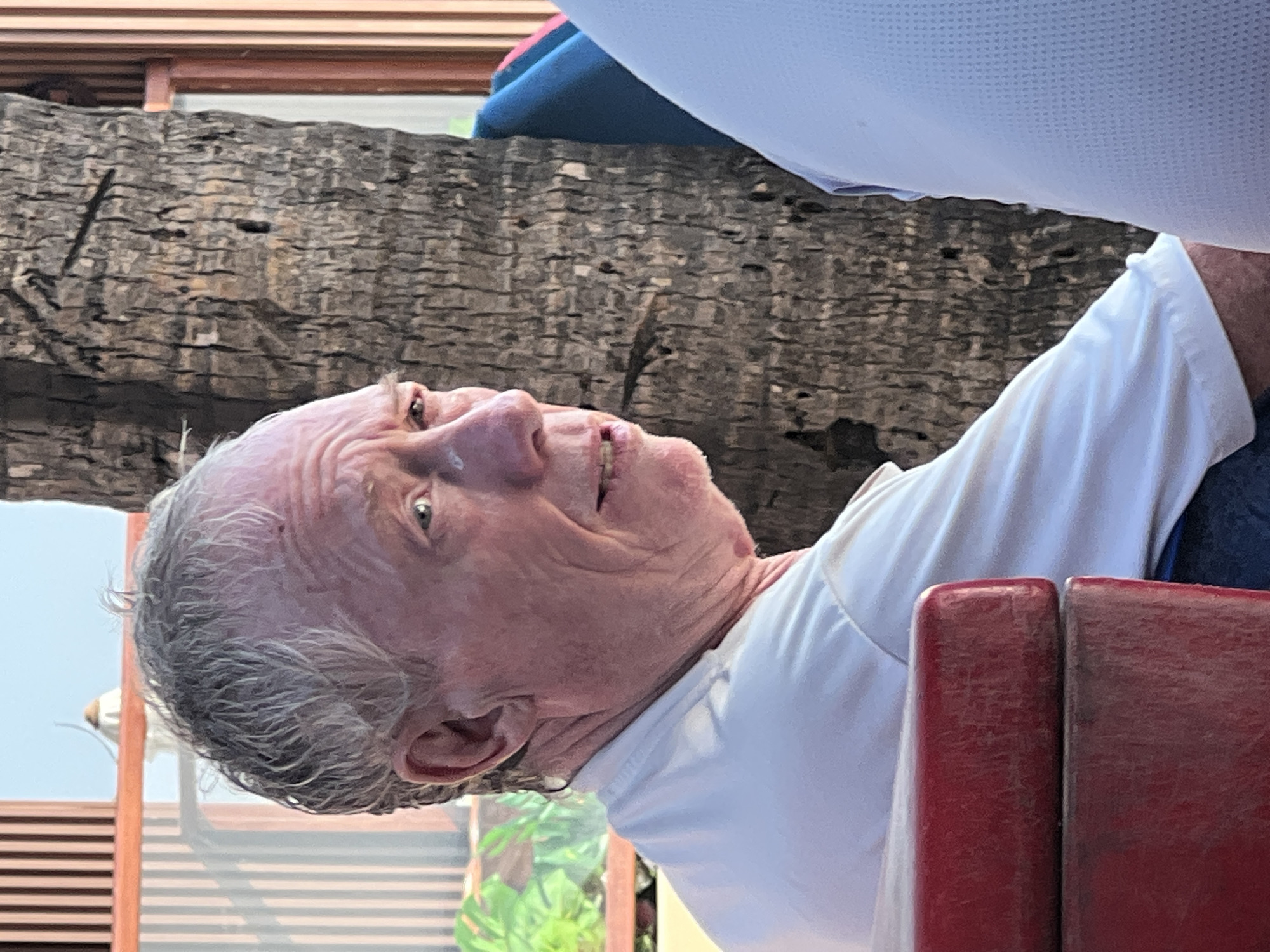 A close-up, medium shot shows an elderly Caucasian male with thinning grey hair, pale skin, and deep wrinkles, facing right. He is wearing a white t-shirt and appears to be sitting outdoors, possibly at a table. The background features a large, rough-barked tree trunk, vertical wooden slats, and some greenery, suggesting a tropical or coastal setting. The lighting indicates it is daytime, possibly late afternoon given the warm tones. The man's facial expression is neutral, with his mouth slightly open. Part of another person in a light blue shirt is visible on the right side of the frame, and a red object, possibly part of a chair or table, is in the lower left corner. The scene is likely in Kuta, Indonesia, a popular tourist destination known for its beaches and tropical environment.