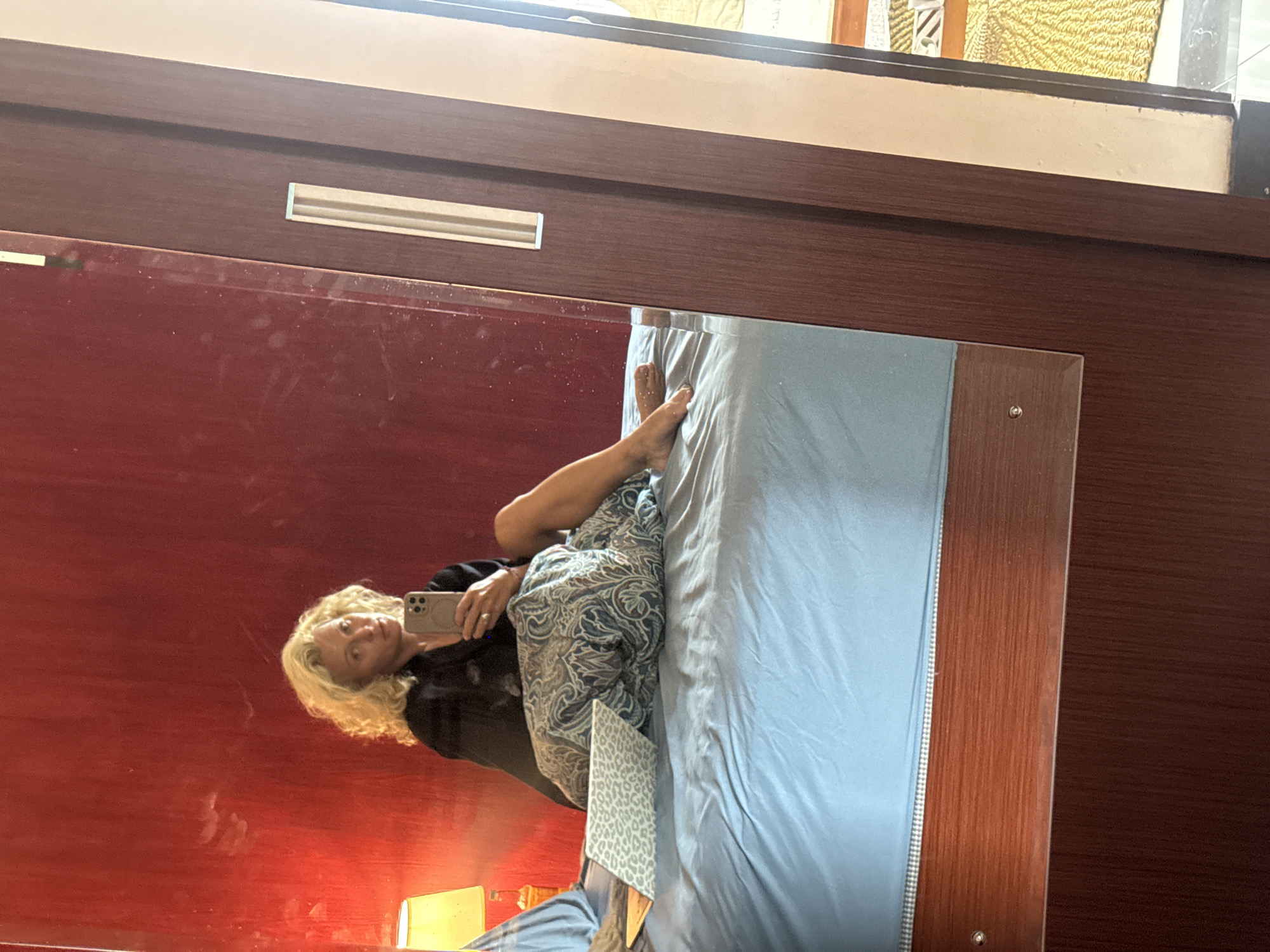 A woman with curly blonde hair is sitting on a bed in a room with a red wall. She is wearing a black top and is holding a phone in her right hand, appearing to take a selfie. A laptop with a leopard-print case is visible on the bed next to her, partially covered by a patterned blanket. The bed has light blue sheets. To the right of the woman, a large wooden wardrobe with a mirrored door is visible, reflecting part of the room. A yellow lampshade can be seen on the left side of the image, against the red wall. The floor appears to be tiled or made of stone.
