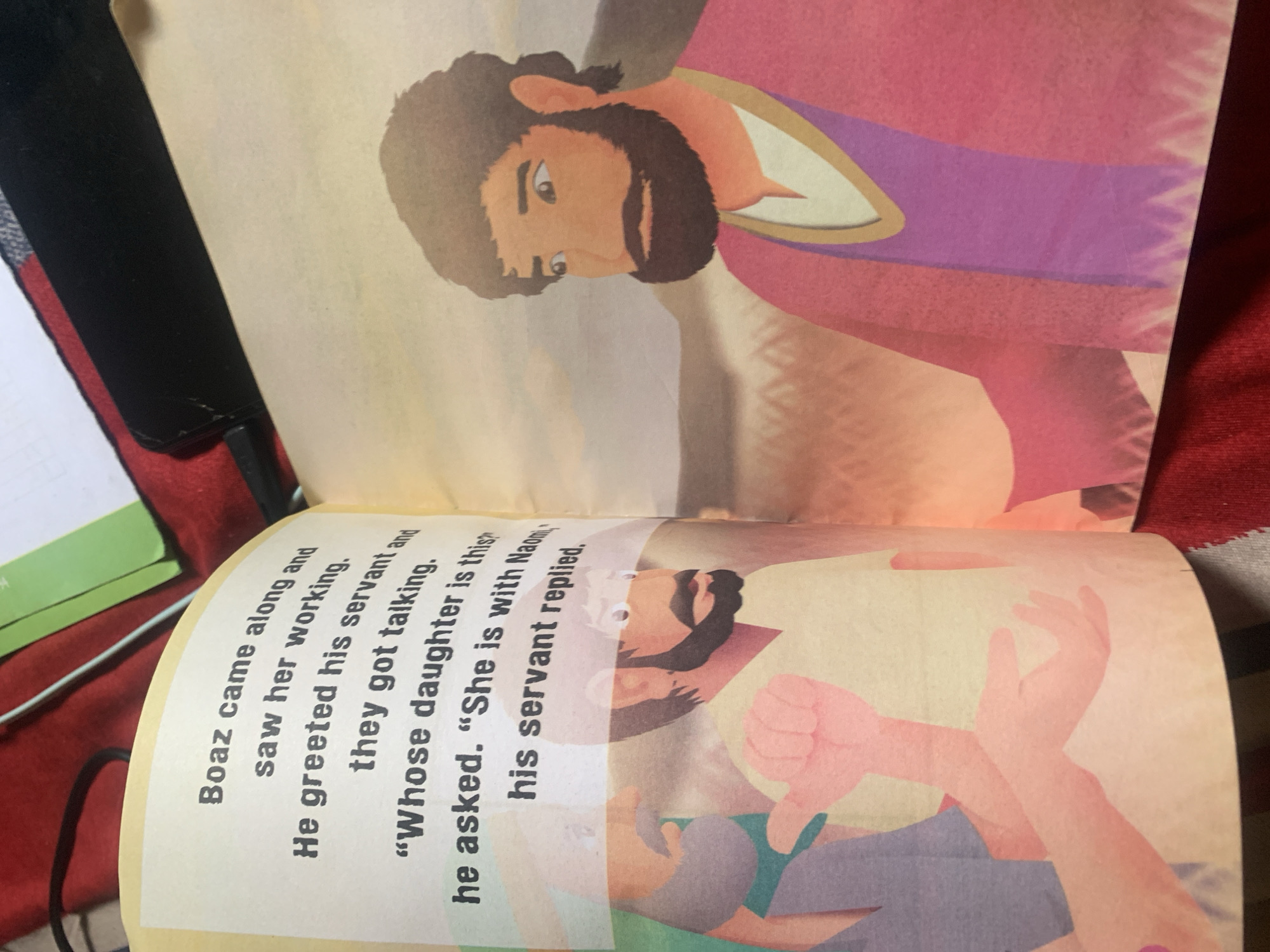 The image depicts an open book with text and illustrations on facing pages. The left page contains black text that reads: "Boaz came along and saw her working. He greeted his servant and they got talking. "Whose daughter is this?" he asked. "She is with Naomi," his servant replied." Below the text are two cartoonish figures. One figure, on the far left, is partially visible, wearing a blue garment and with a purple object in its hand. The other figure, in the center, is more fully depicted, wearing a tan shirt and looking to its left with a concerned expression. The right page features a larger illustration of a man with dark hair and a beard, wearing a pink robe over a purple garment, looking off to the right. The background of both pages appears to be a simplified landscape with muted colors, possibly a field of wheat or grass under a cloudy sky. The image is captured from a slightly elevated angle, showing the red fabric of what appears to be a book cover or blanket beneath the open book. The text and illustrations suggest a narrative from a storybook, likely a religious or historical one. There is no information about Jauro Yinu, Nigeria, present in the image.