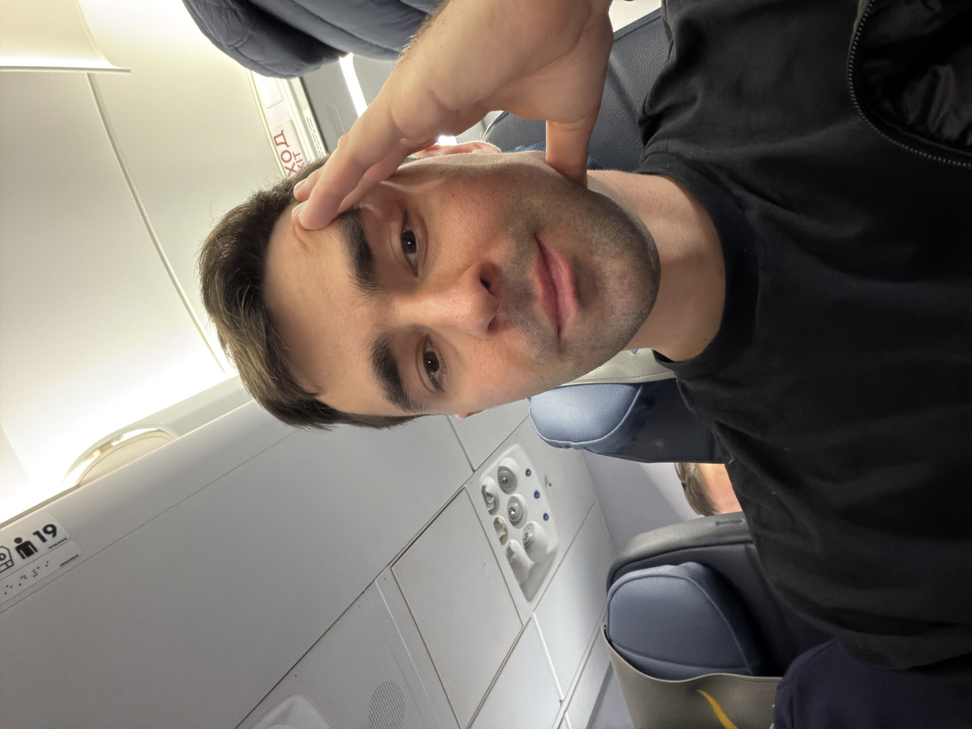 A man is seated on an airplane. He is wearing a black t-shirt and appears to be touching his forehead with his hand. The airplane interior is visible, including overhead controls and seat rows. There is no information in the photo indicating that the location is Tbilisi, Georgia.