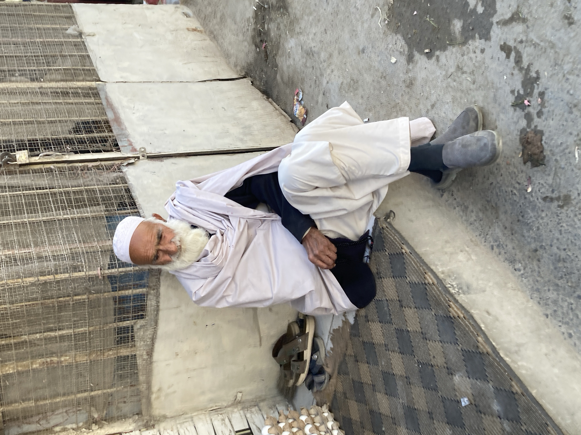 An elderly man with a white beard and a white cap is lying on a patterned mat next to a building in what is likely Quetta City Tehsil, Pakistan. He is wearing a white robe over a black shirt and black socks with grey shoes. Next to him are a pair of sandals and a carton of eggs. The man is resting near a textured concrete surface, possibly a building foundation or sidewalk. The environment appears to be a street or alley.