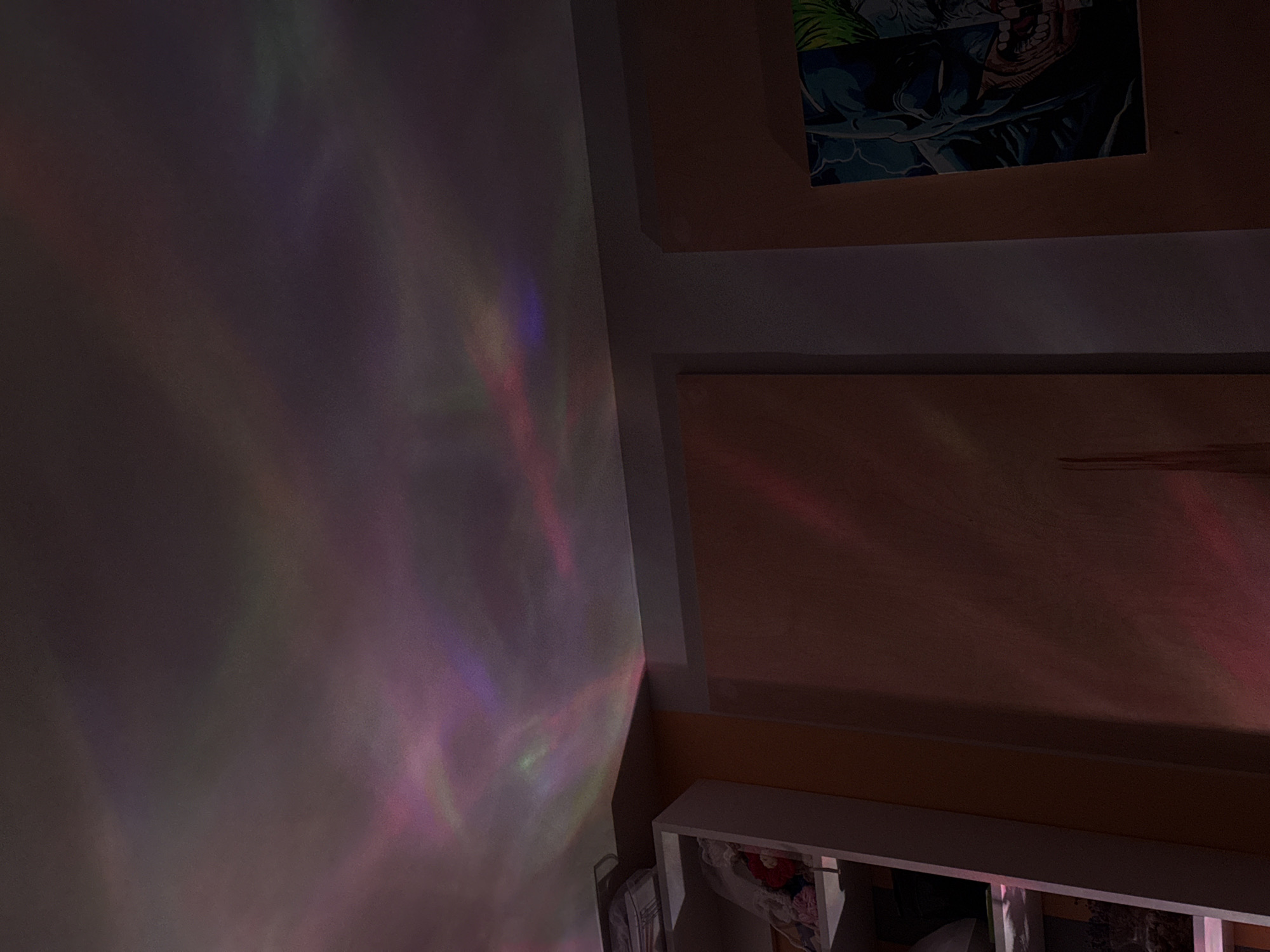 The image shows a section of a room with light projections on the wall. On the right side, there is wooden furniture with a white shelf beneath. The shelf contains various items. Above the wooden furniture, a framed picture is visible. The left wall displays colorful light patterns, which appear to be projected. The overall lighting in the room is dim, enhancing the visibility of the projections. The image does not show any indication of the city of Perm, Russia.