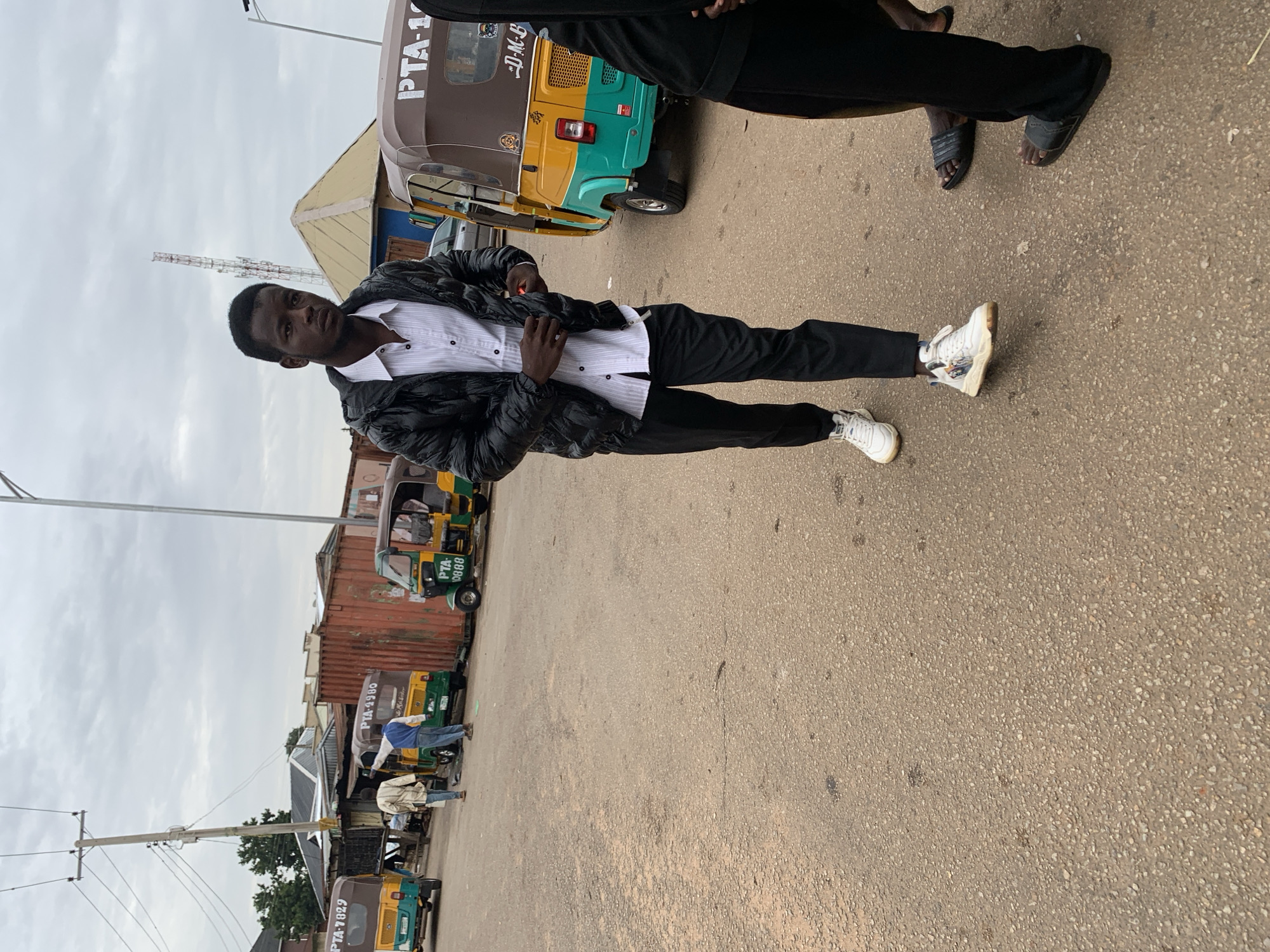A young man wearing a black puffer jacket, a white striped shirt, and black pants walks on a paved street in Jos, Nigeria. He is wearing white sneakers with colorful designs. Behind him, other men are walking and standing, and several yellow and green auto-rickshaws, commonly known as "Keke Napep," are parked on the side of the road. One auto-rickshaw is labeled "PTA 0088," and another has "PTA-7829" visible. The sky is overcast, and in the background, there are buildings, shipping containers, and a telecommunications tower. The scene appears to be a busy street or transport hub.
