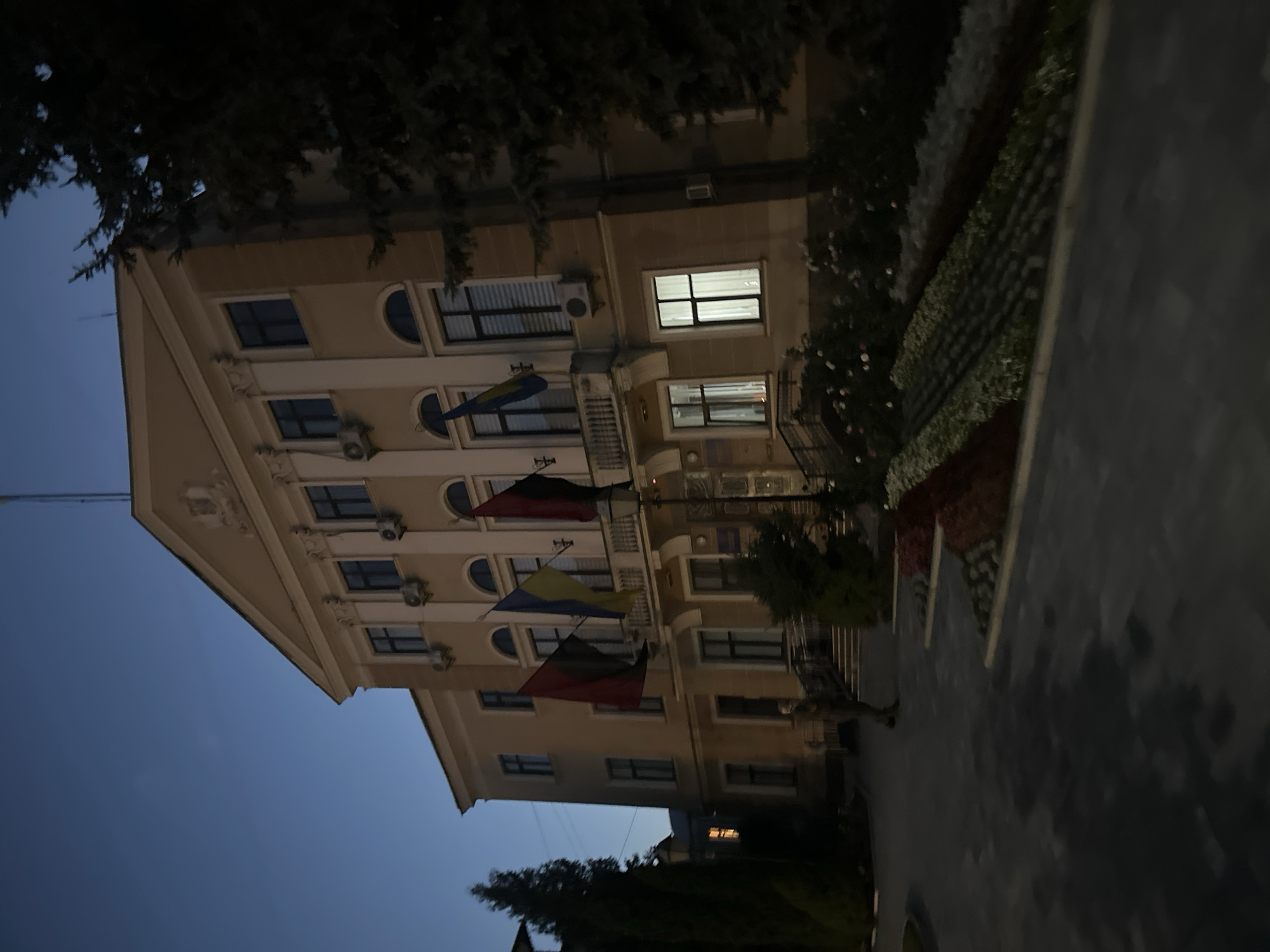 The image shows a building with three flags displayed on its facade. The flags are Ukrainian, and two other flags with black and red colors. The building appears to be a public institution. A person is standing on the sidewalk in front of the building. The image was taken at dusk. The building is located in Ternopil, Ukraine, and appears to be the Ternopil City Council building.