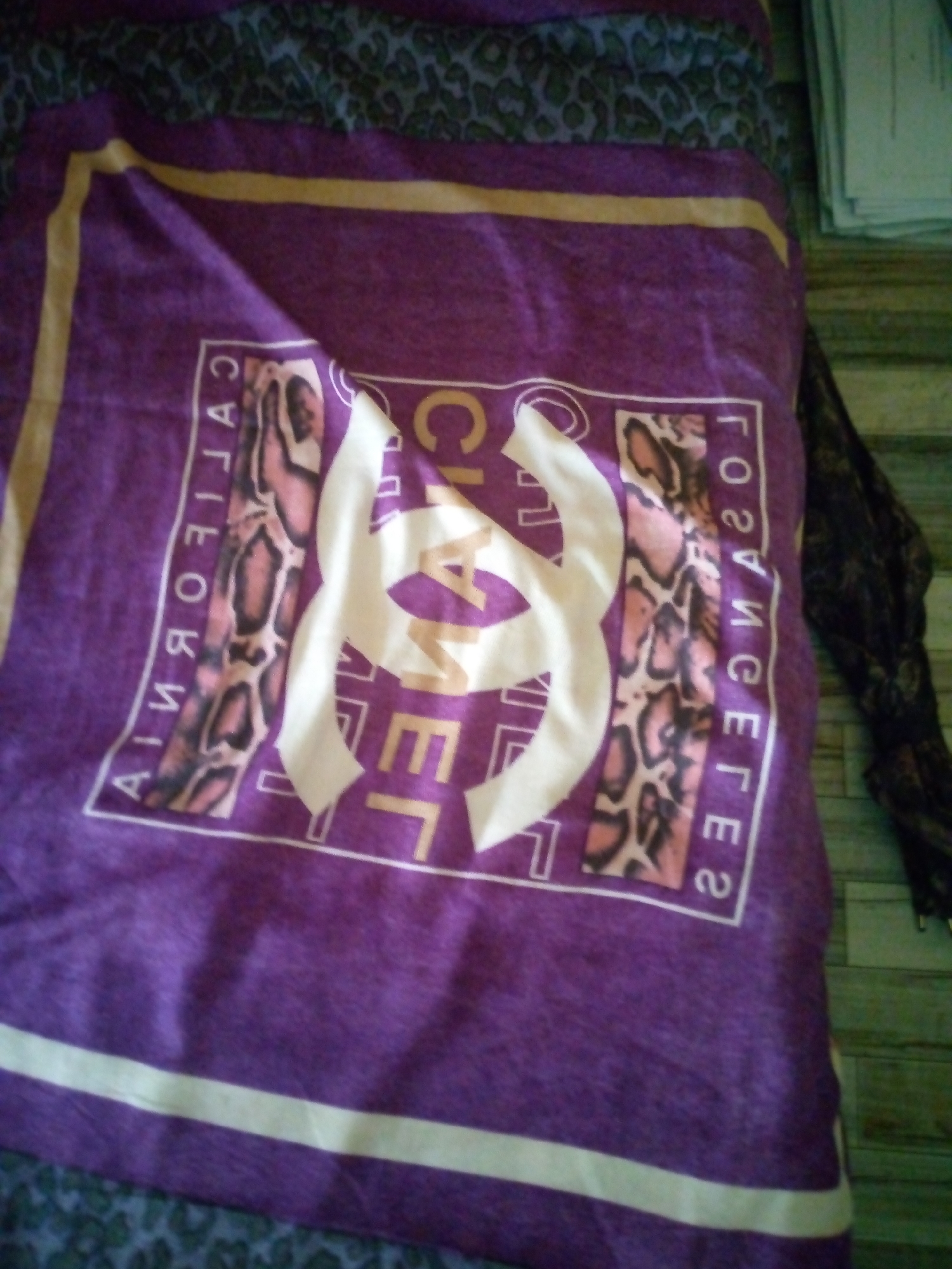 The image shows a close-up of a purple blanket or fabric with a yellow border. In the center of the fabric is a large, white Chanel logo, consisting of two interlocking C's. Inside the logo, there are rectangular insets containing images of what appear to be snakeskin patterns and possibly faces or figures. Along the left edge of the fabric, the word "CALIFORNIA" is written vertically in white capital letters, with the snakeskin pattern interspersed within the letters. Along the right edge, the words "LOS ANGELES" are written vertically in white capital letters, also with the snakeskin pattern.
The fabric is draped over what looks like another patterned material, possibly a bedspread or blanket with a leopard print in shades of grey and dark green. To the right of the purple fabric, a dark, lacy material is visible, suggesting it might be part of clothing or another decorative item. The background appears to be a floor with a wood-like texture, possibly linoleum or tiled flooring. The lighting suggests it's an indoor setting, but the time of day and weather are not discernible. No specific activity or event is taking place, and the image focuses solely on the textile with its prominent branding.
