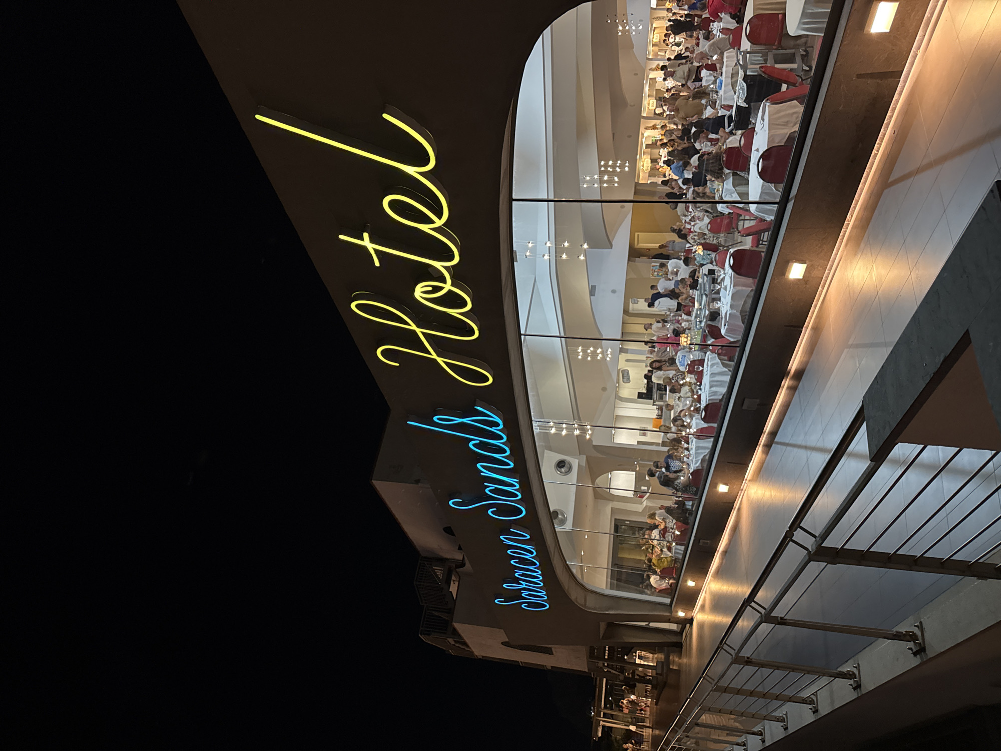 The image shows the "Saracen Sands Hotel" in Capaci, Italy, at night. A neon sign with blue lettering spelling "Saracen Sands" is visible on the side of the building, above a large glass window. To the right of this sign, a yellow neon sign spells "Hotel". Inside the hotel, through the window, a restaurant is illuminated, with numerous tables covered in white tablecloths and red chairs. People are seated at these tables, some appearing to be dining and others socializing. The scene is set against a dark night sky. A metal railing and a walkway are in the foreground, suggesting an outdoor area or balcony overlooking the restaurant. The overall impression is of a lively evening at a hotel and restaurant.
