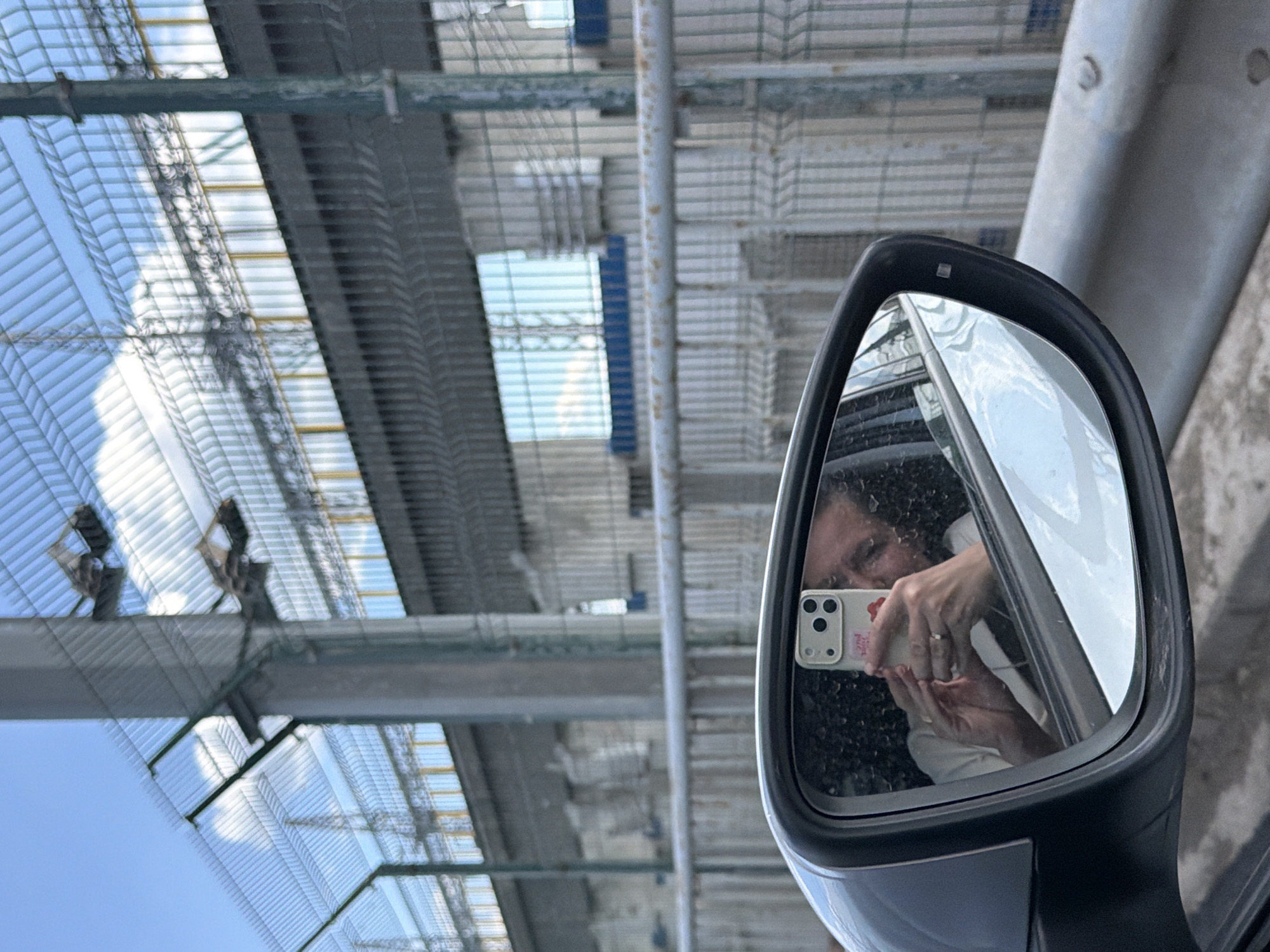 In the foreground, a car's side mirror reflects a person taking a selfie with a smartphone. The person is wearing a light-colored shirt and jewelry. In the background, a large metal fence stretches across the frame, with a cloudy sky visible above. The fence appears to enclose an industrial area with concrete structures and more fencing. The setting is likely Chaikovsky, Russia, given the presence of a large industrial complex and fence. The overall scene suggests a moment captured while traveling through an industrial zone.