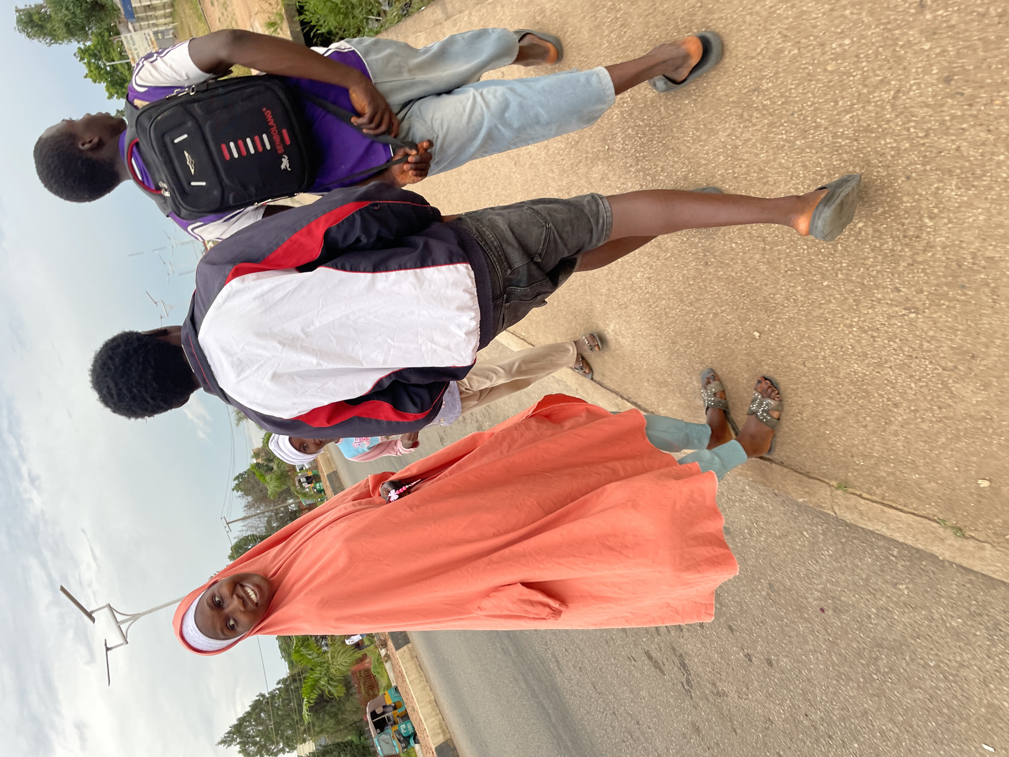 A group of four individuals are walking on a sidewalk next to a road in Jos, Nigeria. The person in the foreground, a woman wearing a bright orange hijab and a matching garment, is smiling and looking to her left. Next to her and slightly behind is a child wearing a white hijab. Further behind them, two young men are walking. One is wearing a purple shirt and a black backpack. The other is wearing a jacket over a white and red shirt, and dark shorts. The road beside the sidewalk has a paved surface with faint white lines. A three-wheeled vehicle, commonly known as a Keke Napep, is parked on the side of the road. The environment suggests an urban setting with buildings and vegetation visible in the background under an overcast sky.