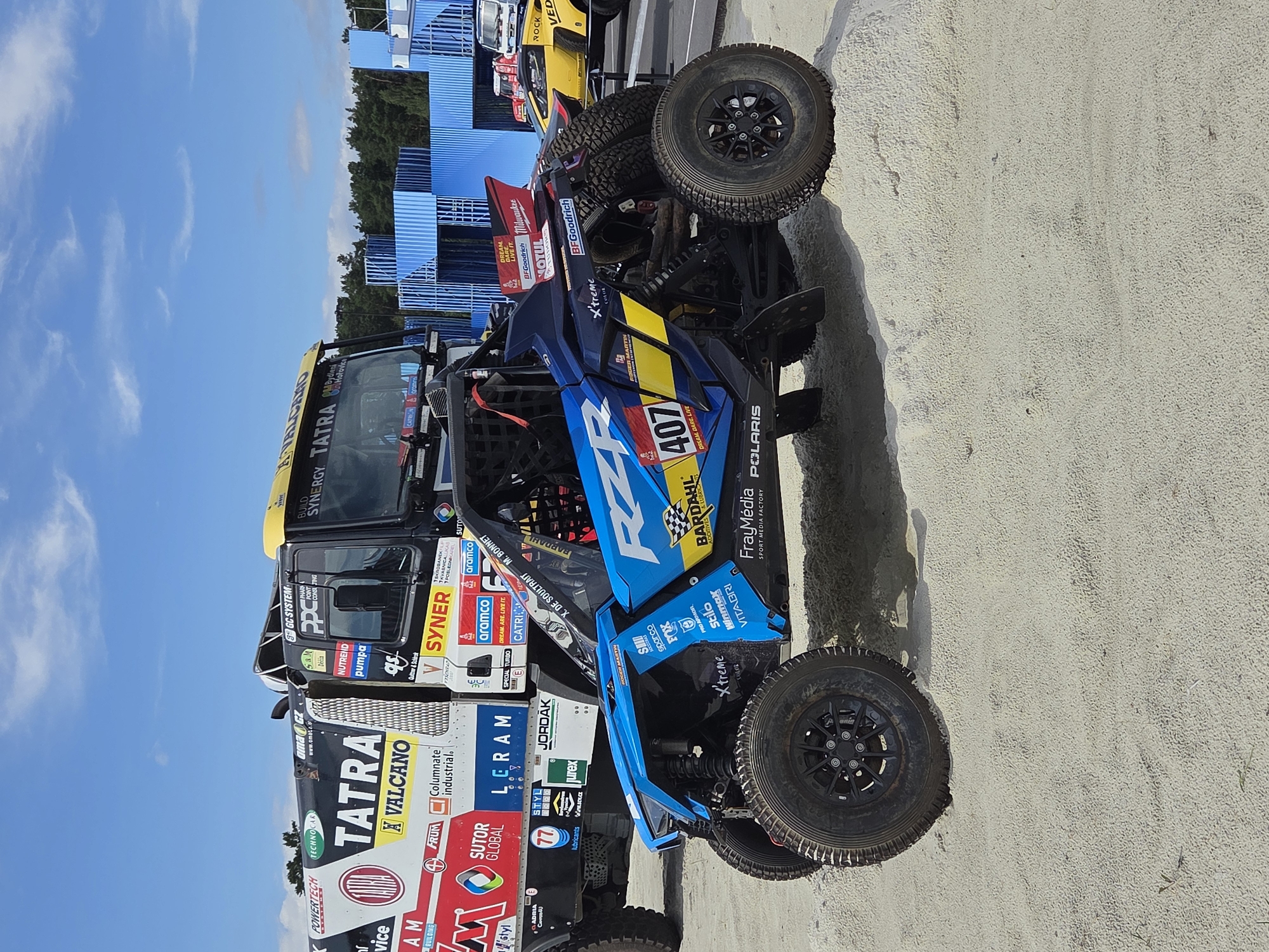 The image depicts an outdoor scene with two off-road vehicles and a truck in the foreground. The primary subject is a blue and black buggy with the number "407" prominently displayed on its side, featuring sponsor logos such as "RZR," "Bardahl," and "Polaris." To the left of the buggy is a large white and black truck with the "Tatra" logo and various other sponsor decals. In the background, there are blue shipping containers, a forest, and a clear blue sky with some clouds. The ground is covered in sand, suggesting a desert or sandy terrain, which is unlikely in Yekaterinburg, Russia. The lighting indicates it is daytime. No people are visible in the image.