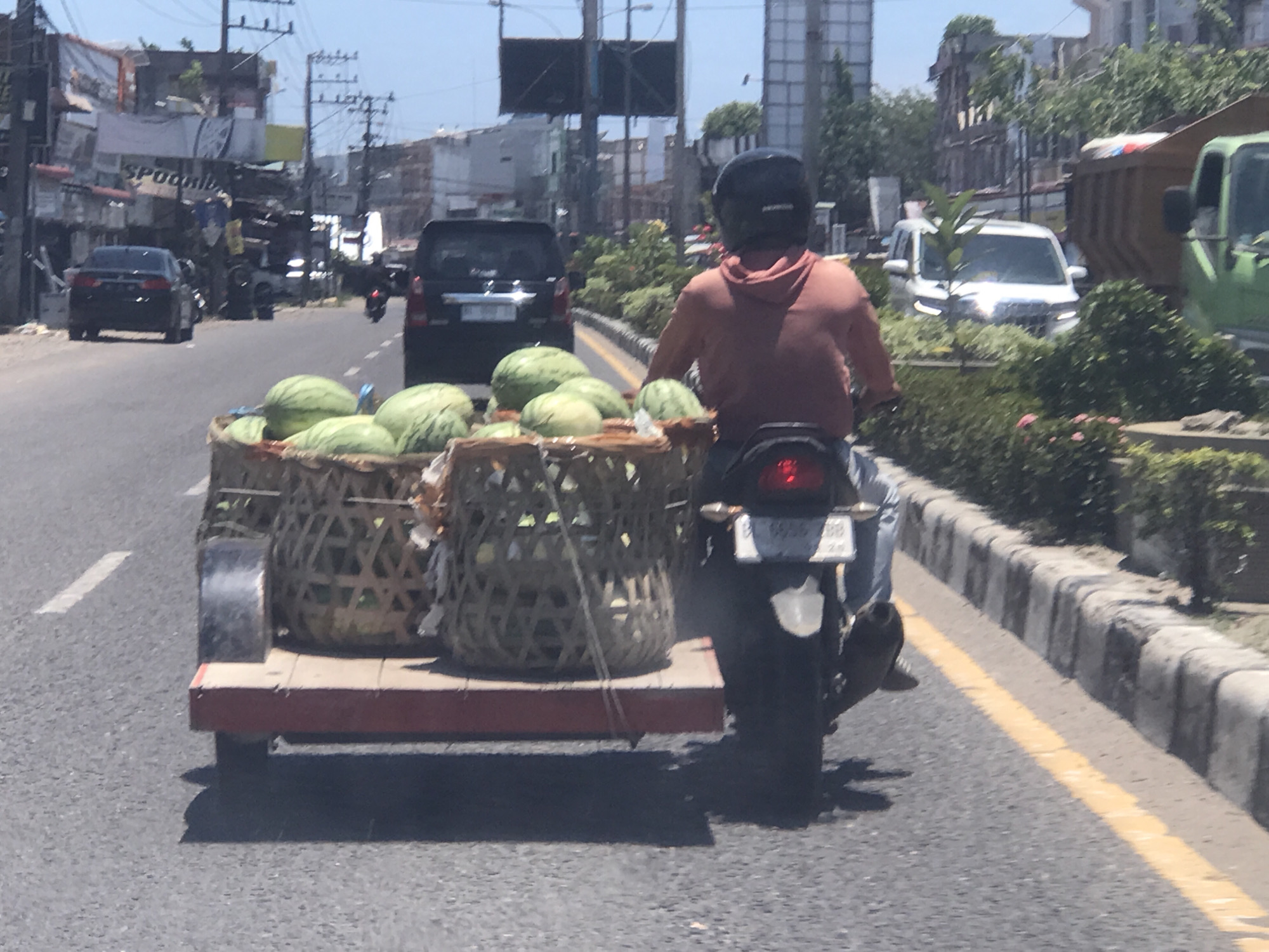 The image shows a man riding a motorcycle with a trailer carrying several watermelons on a road in Lhokseumawe, Indonesia. He is wearing a helmet and a hooded top. The watermelons are in woven baskets on a small wooden trailer. The motorcycle has a license plate that reads "BL 6658 BB". The road has a yellow dividing line and a concrete curb on the right side. Other vehicles, including cars and trucks, are visible in the background along with shops and buildings. The weather appears to be sunny.