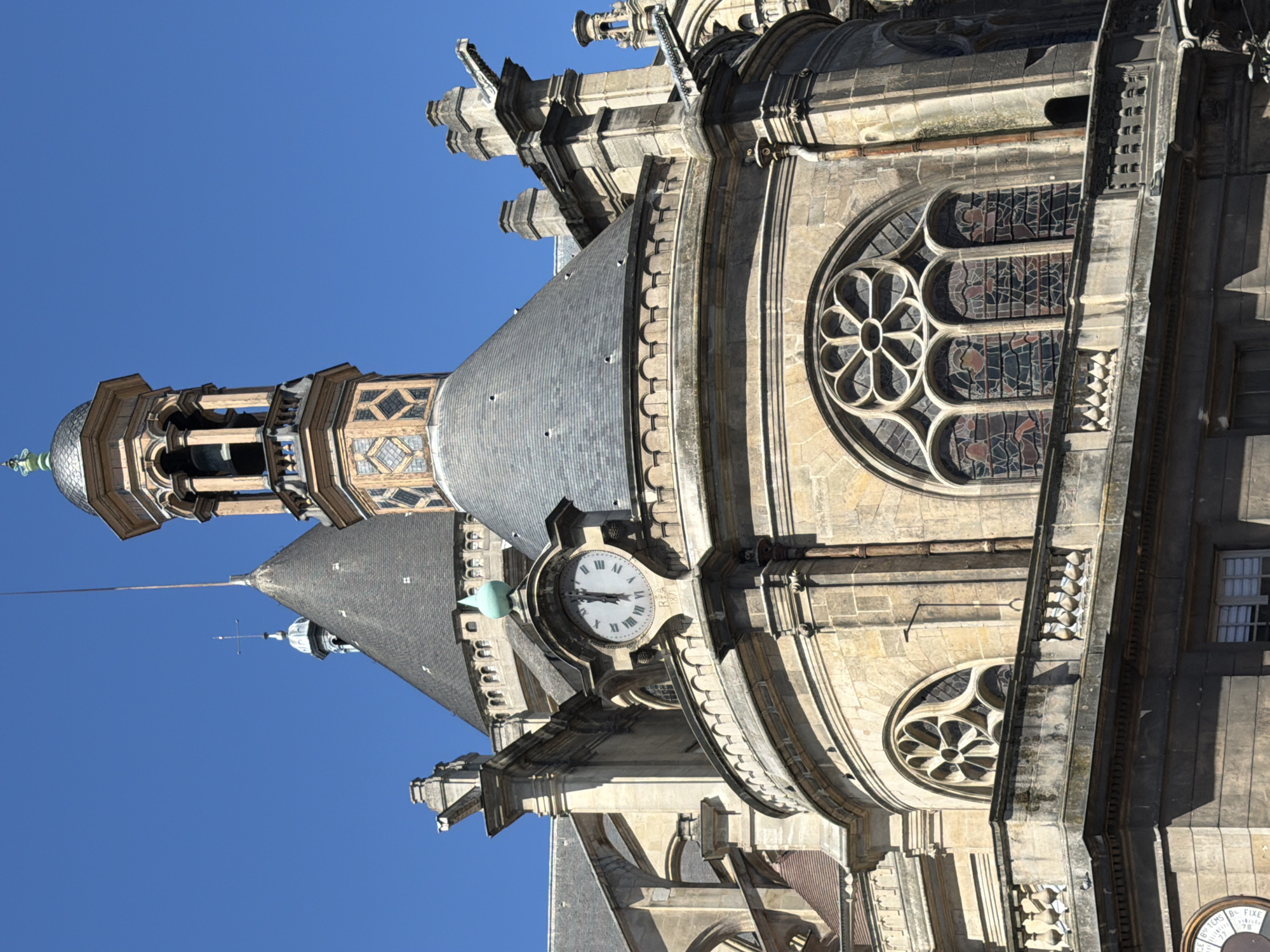 The image shows the facade of a historic stone building, likely a church or cathedral, under a clear blue sky. The architecture features a prominent clock tower with a bell, ornate stonework, arched windows with stained glass, and gargoyles. The clock face, with Roman numerals, is visible and appears to read approximately 1:49. The building's materials are weathered stone and grey slate roofing. The scene suggests a daytime view in an urban setting, with no people or active events depicted. The architectural style is consistent with Gothic or Neo-Gothic design. This is likely the Sainte-Chapelle, a royal chapel in the Gothic style on the Île de la Cité in Paris, France.