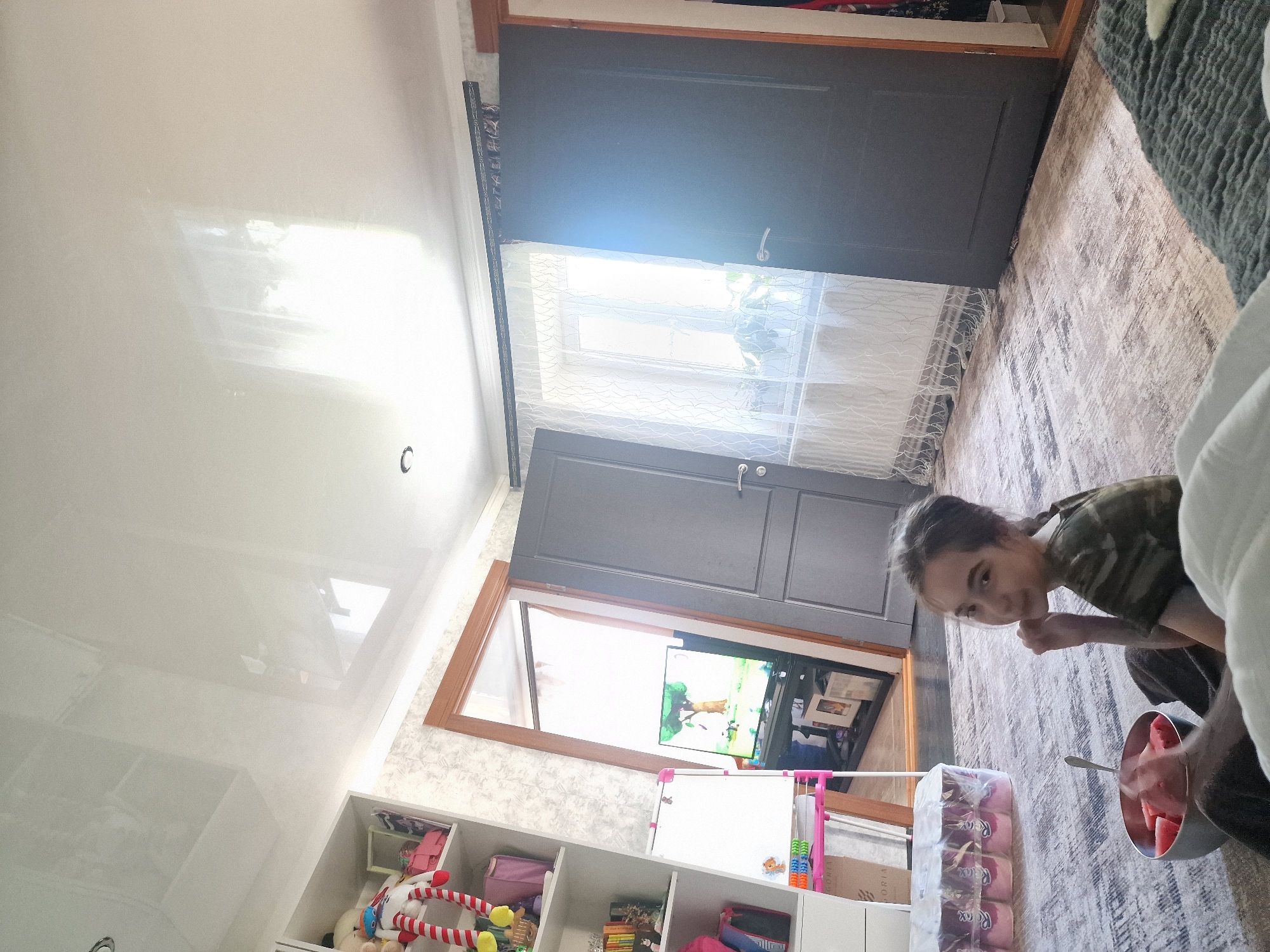 A young person with dark hair is sitting on a patterned rug in a room. They are wearing a camouflage t-shirt and dark pants. The person is looking at the camera and holding a bowl of sliced watermelon with a spoon. The room appears to be a living space, with furniture and shelves visible in the background. There is a closed dark gray door and an open doorway leading to another area with a television playing. The ceiling is white and glossy, reflecting light. The overall lighting suggests it could be daytime. The presence of toys on shelves indicates a child-friendly environment. The setting is likely a home in Kursavka, Russia.
