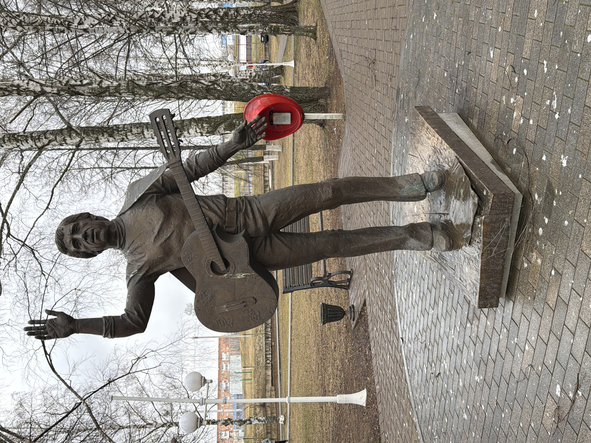 A bronze statue of a male figure stands prominently in an outdoor public space in Votkinsk, Russia. The statue depicts a musician or bard, holding an acoustic guitar across his body. He is dressed in a long-sleeved shirt and trousers, with his head tilted upwards and mouth open as if singing or speaking. His right hand holds a bright red, bowl-shaped object, possibly a hat or basin, inside which a small, light-colored object resembling a telephone receiver is visible. His left arm is extended outwards, palm open.
The statue rests on a dark, rectangular stone base. The surrounding ground is paved with grey interlocking stones, which are notably marked with numerous white splatters, likely bird droppings. In the background, several birch trees with bare branches suggest a late autumn, winter, or early spring season. The sky is overcast, providing soft, diffused light.
To the left of the statue, two decorative lampposts with globe lights are visible. Behind the statue, a dark park bench is partially seen, and further in the distance, a multi-story building with a reddish-brown facade and numerous windows indicates an urban or suburban setting. Patches of dry, brown grass or dirt are interspersed among the paved areas and trees. No clearly legible text is visible in the image.