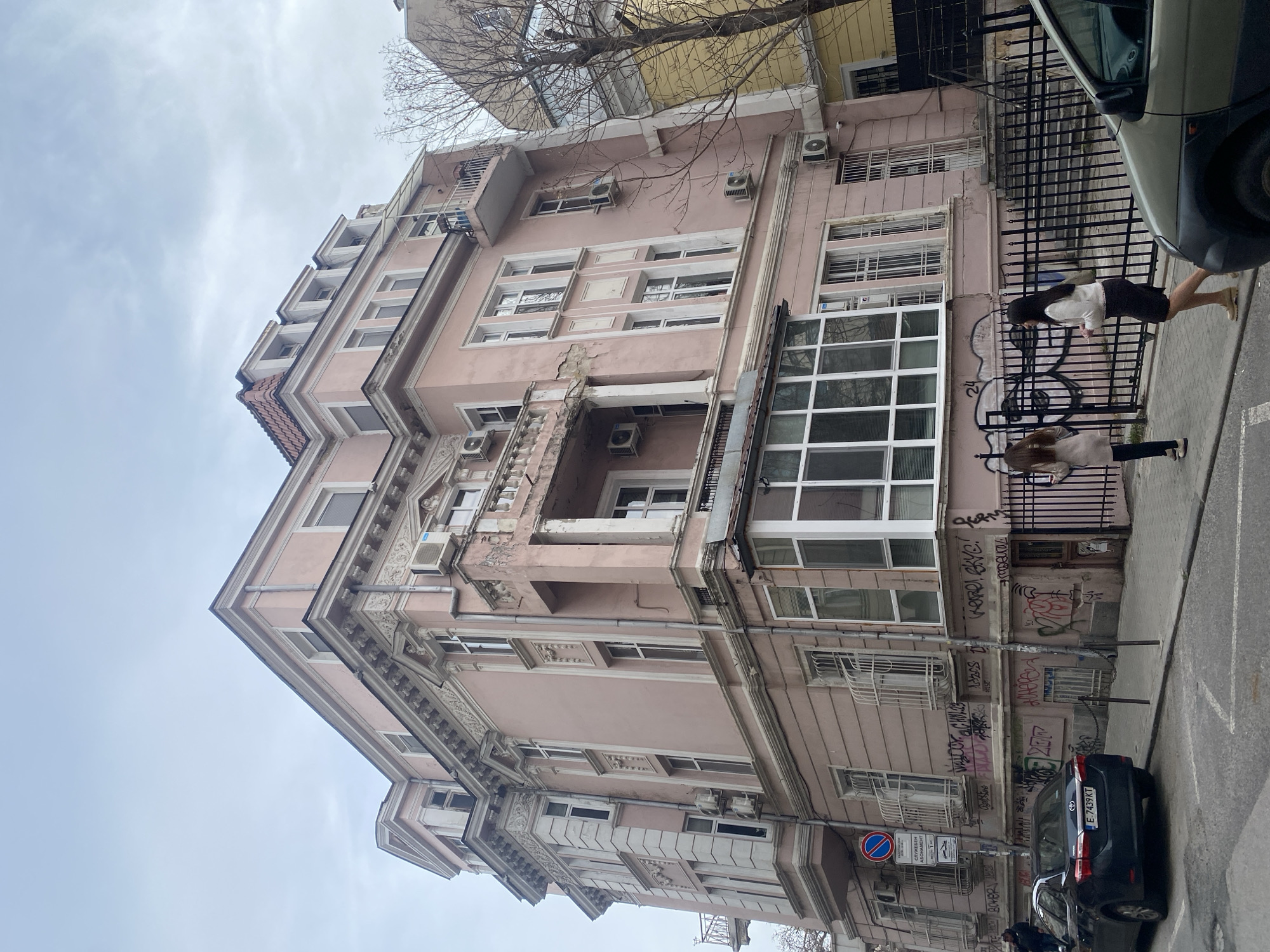 The image depicts a street scene in Sofia, Bulgaria, featuring a pink, multi-story building with ornate architectural details. The building shows signs of age and some disrepair. It has multiple windows, some of which have air conditioning units installed. There's a black metal fence along the sidewalk, partially covered in graffiti. Two women are walking on the sidewalk next to the fence. A dark-colored car is parked along the curb. Street signs are visible. The sky is overcast.
