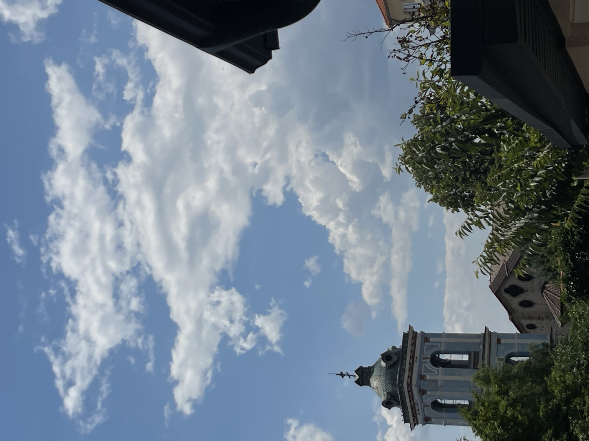The image depicts a view of the town of Karlovo, Bulgaria, on a cloudy day. In the foreground, a dark, angled roofline partially obstructs the view. To the left, a tall, blue bell tower with arched windows stands prominently, topped with a green, multi-tiered spire and a cross. Adjacent to the tower, part of a building with a stone facade and dark arched windows is visible. Lush green trees and foliage surround the buildings, with some bare branches of a tree visible on the right. The sky above is a bright blue, filled with dramatic, white cumulus clouds. The scene captures an architectural and natural landscape under a dynamic sky.