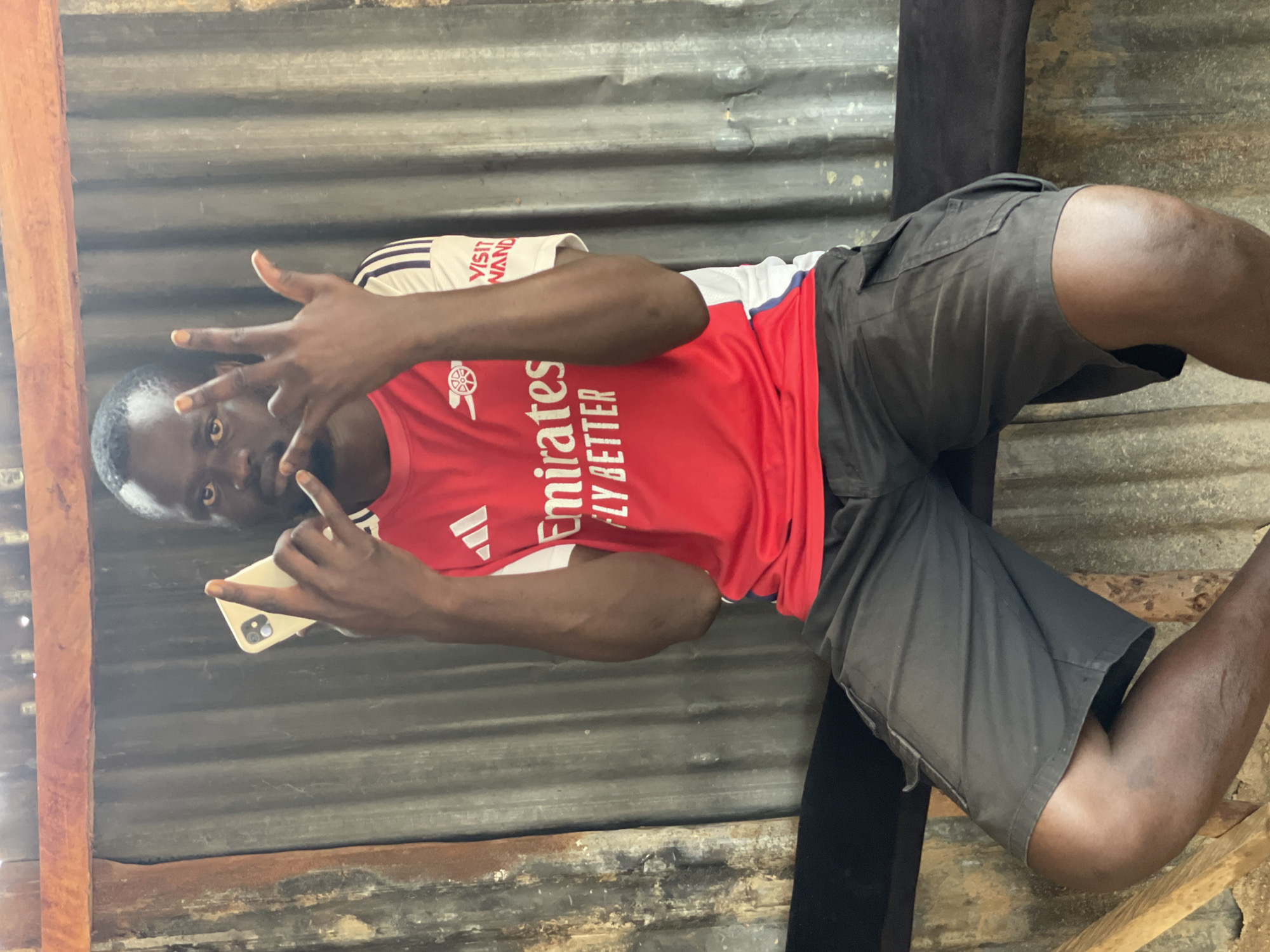 A Black man is seated and looking at the camera, holding a smartphone in his left hand and making a hand gesture with his right hand. He is wearing a red Arsenal soccer jersey with white trim and gray cargo shorts. The background appears to be a corrugated metal wall, with wooden beams above. The setting seems rustic and possibly outdoors or in a shed-like structure. There is no identifiable location information within the image itself, and therefore no specific mention of Durimi, Nigeria can be made.