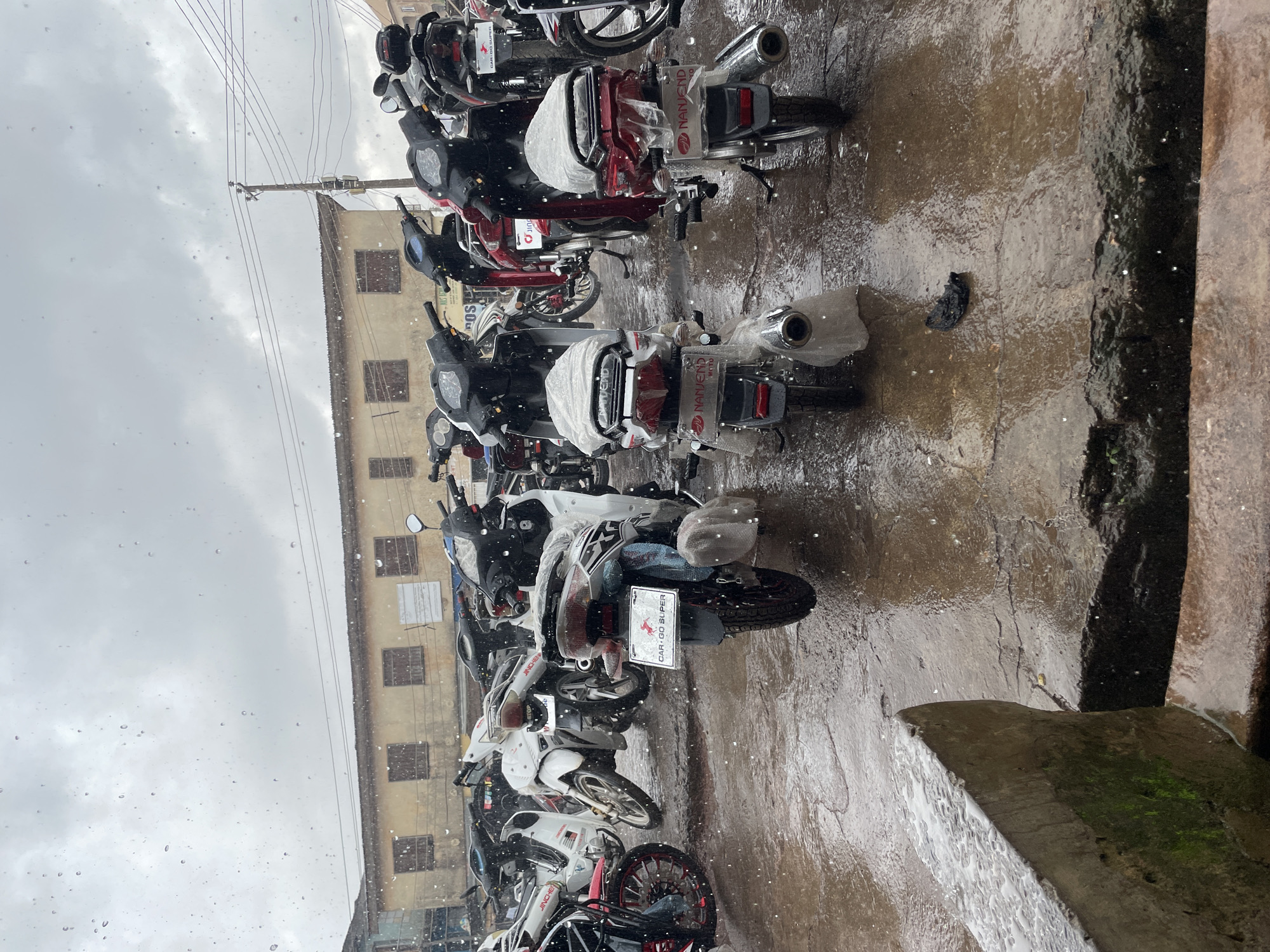 The image depicts a street scene in Jos, Nigeria, under heavy rainfall. A row of motorcycles, primarily scooters and smaller displacement bikes, are parked on a wet, cracked asphalt surface. The bikes are of various colors, including white, black, and red, and some are partially covered in plastic sheeting. Water is visibly falling and splashing on the ground and the vehicles. In the background, a two-story building with a pale yellow facade and shuttered windows stands under a cloudy, overcast sky. Power lines and a utility pole are also visible. The overall impression is one of a commercial area experiencing inclement weather. There are no people clearly visible in the frame, and the focus is on the parked motorcycles and the wet environment.