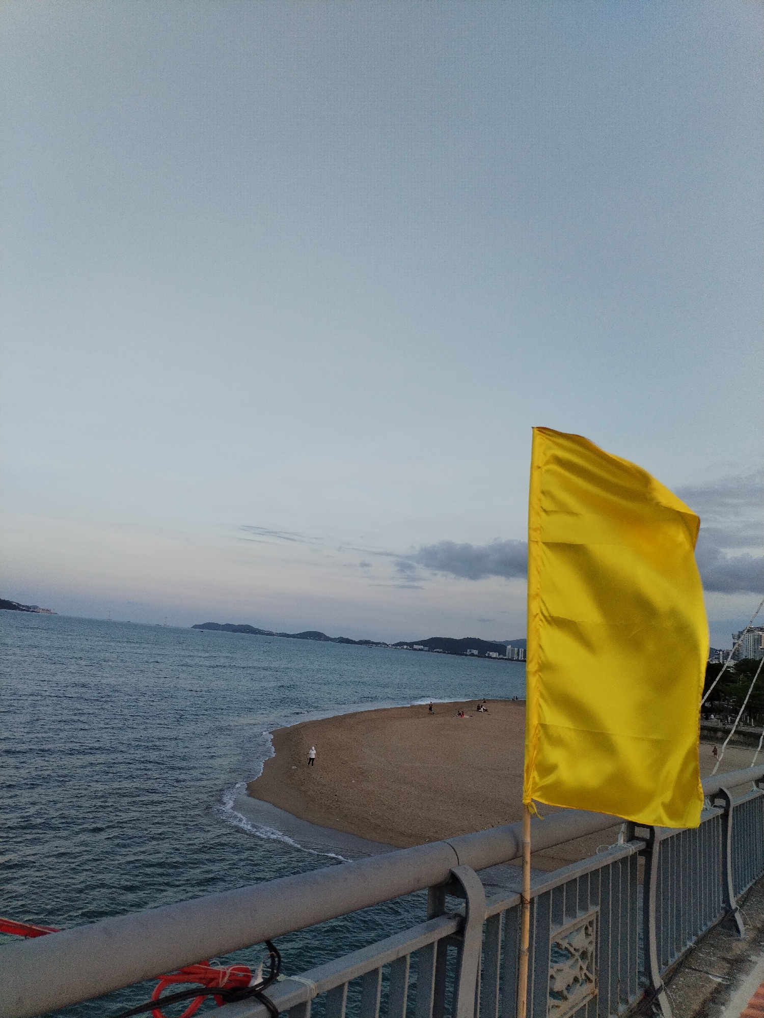 The image shows a coastal scene in Nha Trang Ward, Vietnam. In the foreground, a bright yellow flag is mounted on a grey metal railing. Behind the railing, there is a sandy beach with several people scattered along the shore. The beach meets the ocean, which extends towards distant islands and a skyline with buildings. The sky is overcast with hues of grey and blue.