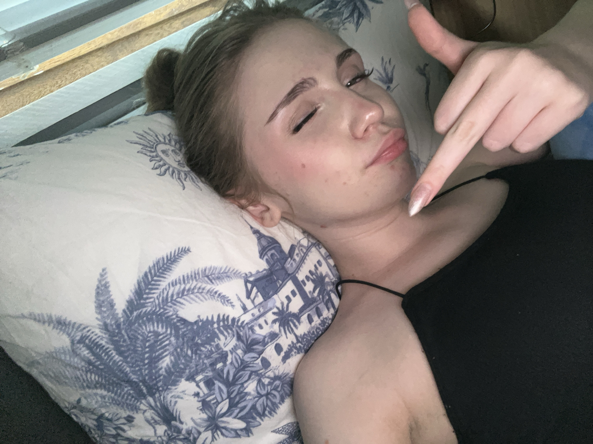 A close-up, high-angle shot shows a young woman lying on a pillow with a blue and white botanical print. Her eyes are closed, and her head is tilted back. A person's hand is in the foreground, with a thumb extended upwards and an index finger pointing towards the woman's chin. The woman is wearing a black tank top. The background consists of horizontal blinds and a wooden surface. There is no visible indication of the city of Ashkadar, Russia.