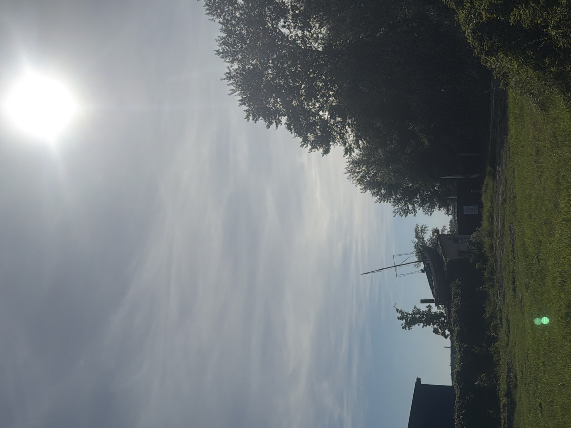 In the city of Vzmorye, Russia, a unique scene unfolds under a bright, sunlit sky. Dominating the frame is a weathered wooden boat, inexplicably positioned atop a building, its mast reaching towards the heavens. The boat appears to be tilted, adding to its surreal placement. Surrounding the building are dense trees and overgrown vegetation, creating a somewhat wild and untamed atmosphere. In the foreground, a grassy field stretches out, its lush green contrasting with the muted tones of the structures and the sky. The overall impression is one of an abandoned or unconventional space, where the ordinary has been decidedly defied. The bright sunlight casts a strong glare across the image, particularly in the upper portion of the sky, hinting at a clear and possibly warm day. No individuals are visible in the frame.