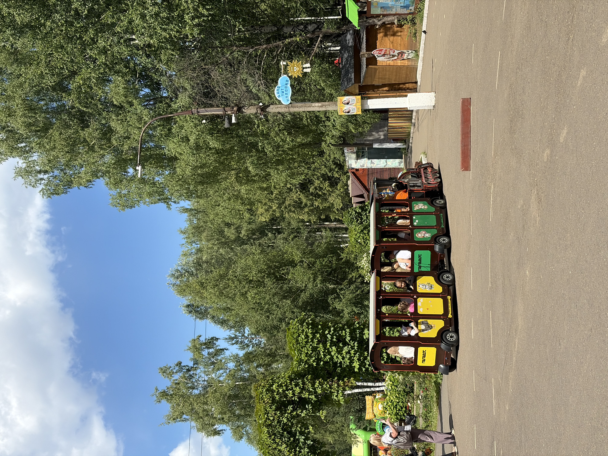 The image captures a bright, sunny day in Chaikovsky, Russia, likely in an amusement park or recreational area. The foreground features an asphalt path with white lane markings. Dominating the mid-ground is a miniature train with several brightly colored carriages, carrying a group of passengers, including children. The train is brown and yellow with green accents and features cartoonish decals on its side. To the left of the train, a woman with a tote bag stands taking a photo. Behind the train, there are small buildings, some with wooden facades, and a section of a fence. Lush green trees and foliage form the background, with a bright blue sky with scattered white clouds overhead. A streetlamp with attached speakers and signage is visible on the right side of the path. The overall impression is one of a pleasant outdoor family outing.