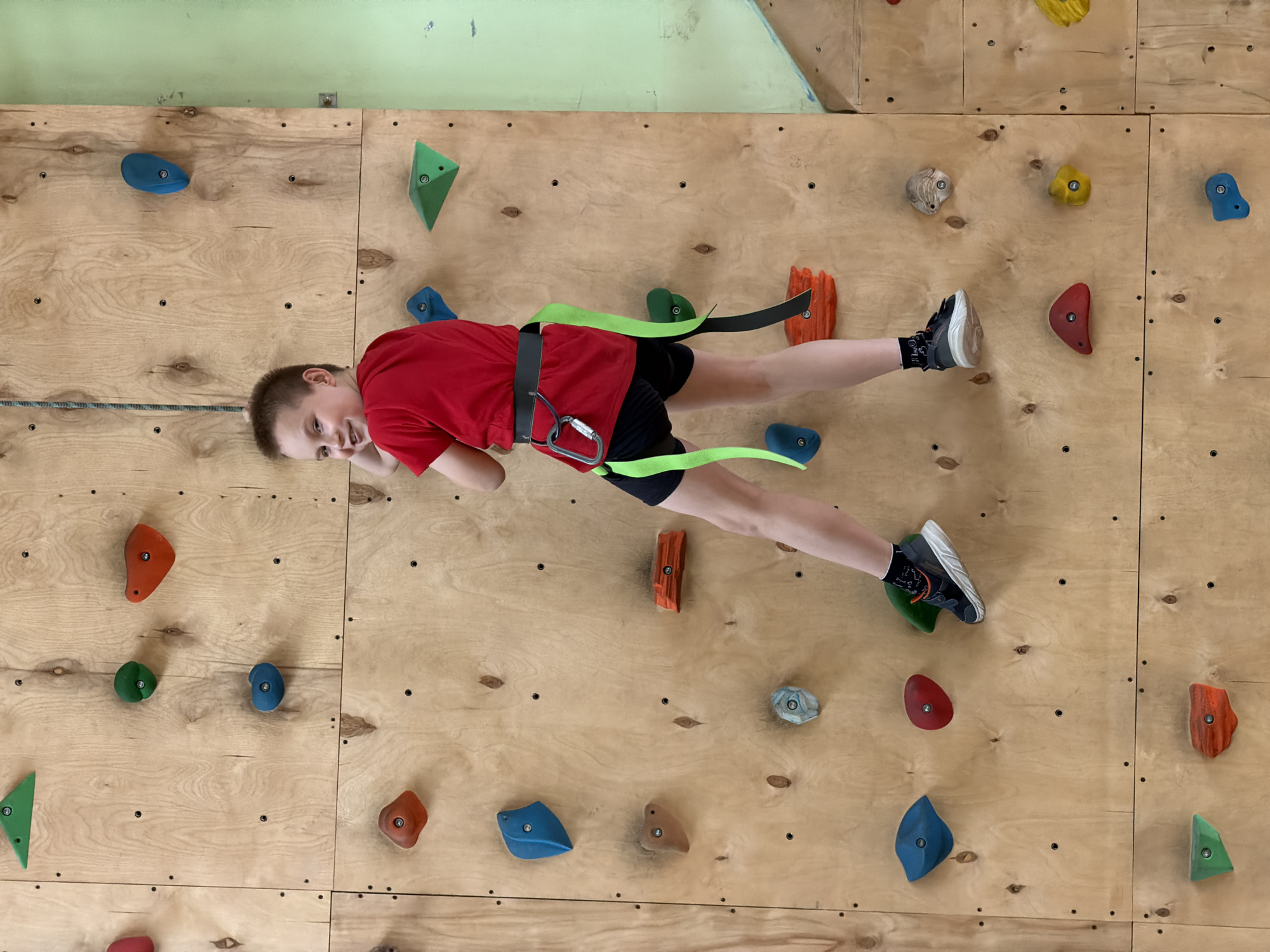 A young boy is captured in a full-body shot, suspended horizontally on an indoor climbing wall. He appears to be around 8-12 years old, dressed in a red short-sleeved t-shirt, black shorts, and black athletic shoes with white soles, paired with patterned black and white socks. A bright green climbing harness with a black strap is secured around his waist and thighs, to which a silver carabiner is attached. A climbing rope, blue and white in color, is visible near his left hip, connected to the harness.
The boy is looking directly at the camera with a cheerful expression, a slight smile on his face, suggesting enjoyment or a playful pause during his climb. His body is stretched out against the wall, showcasing his engagement in the activity.
The climbing wall itself is constructed from numerous light-colored wooden panels, affixed together with visible screws. Scattered across the wooden surface are various colorful climbing holds of different shapes and sizes, including blue, green, red, orange, yellow, and beige. Above the climbing wall, a plain light green painted wall is visible, suggesting an indoor gym or recreation center setting. The lighting appears bright and even, consistent with an indoor environment. No text is visible in the image. The scene conveys a sense of youthful energy and a fun, active experience taking place in Izhevsk, Russia.