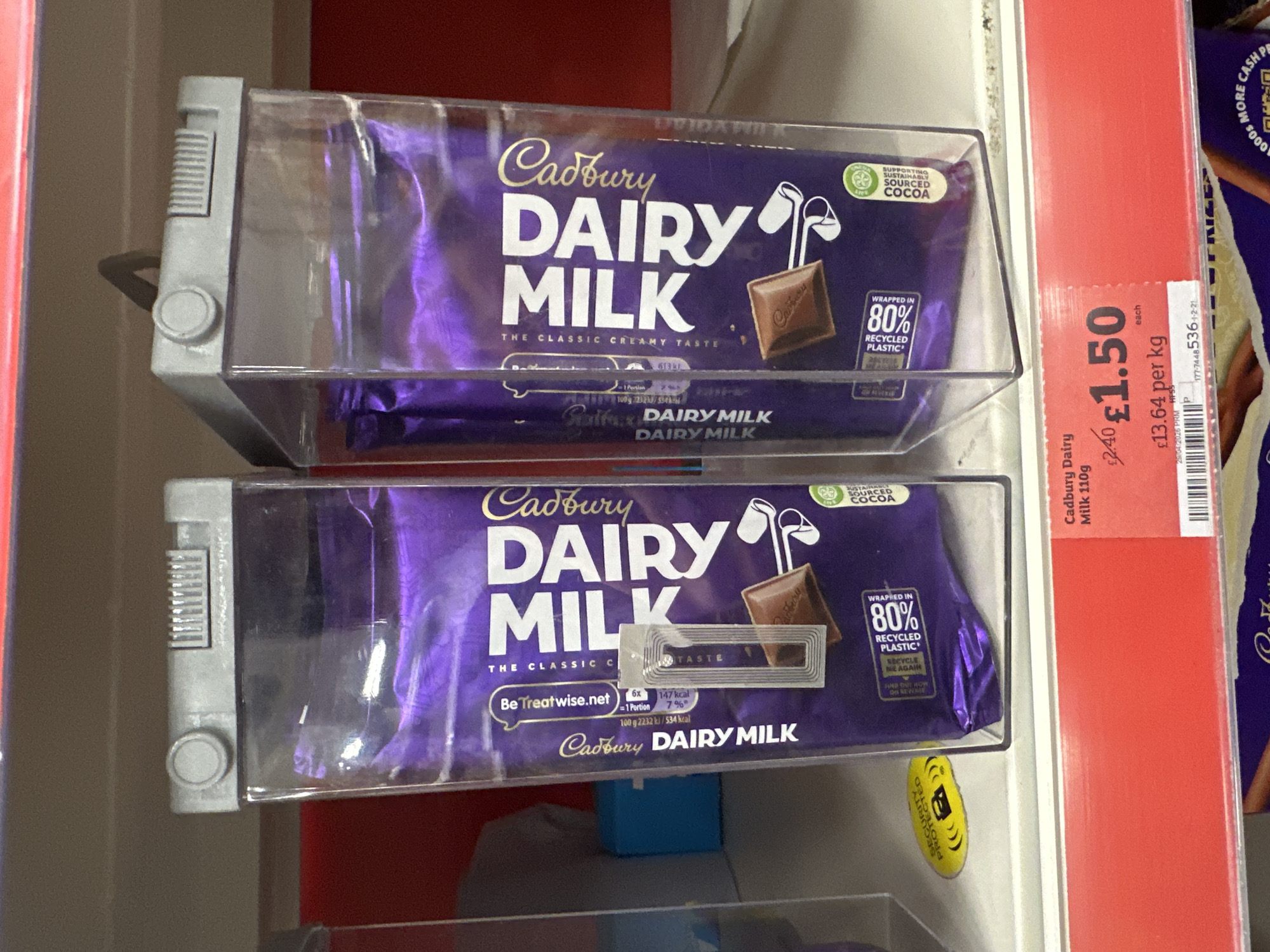 This image shows two bars of Cadbury Dairy Milk chocolate in a refrigerated display case in what appears to be a retail environment. The chocolate bars are in purple packaging with the Cadbury logo and "Dairy Milk" prominently displayed. The packaging highlights that the cocoa is sustainably sourced and the wrapper is made of 80% recycled plastic. A price label next to the chocolate indicates the price is £1.50 per bar, which equals to £13.64 per kg, the price label also includes the date 29/04/2026 and a bar code. There is a security tag attached to the lower bar. The location is possibly in London, United Kingdom.