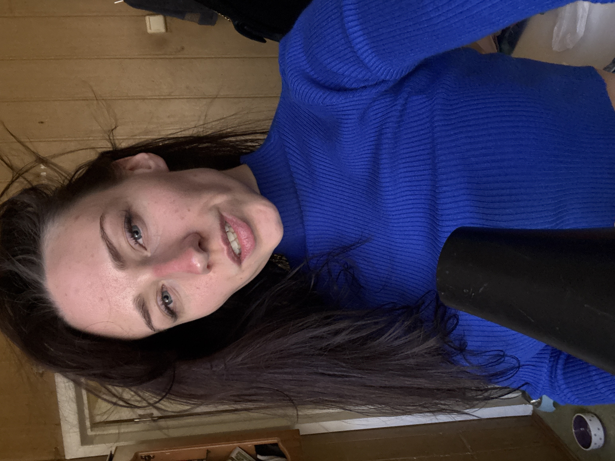 A woman with dark hair and blue eyes is looking at the camera with a slight smile. She is wearing a blue ribbed sweater. She appears to be using a black hair dryer. The background consists of wood paneling and a lower section with what appears to be a cupboard or shelf. The location in Saint Petersburg, Russia, is unknown and cannot be determined from this image.