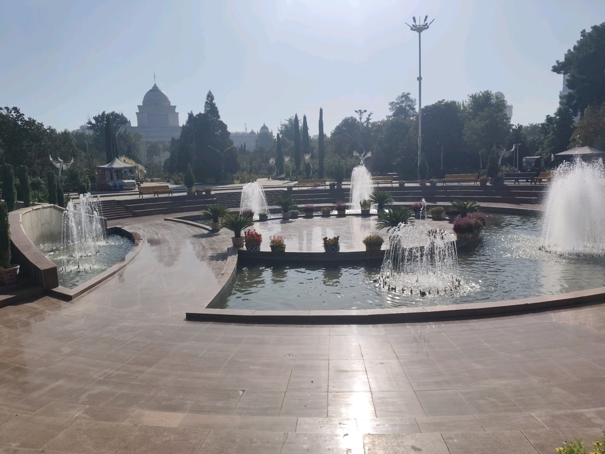 The image depicts a large, well-maintained park in Dushanbe, Tajikistan. The foreground features a polished, light brown tiled area surrounding a multi-tiered fountain system. The central fountain is the largest, with water spraying upwards in a wide arc, surrounded by smaller jets. Several other smaller fountains are visible, some with intricate spray patterns and others forming delicate cascades. The water in the pools reflects the sky and surrounding greenery.
Several planters with small palm-like plants and flowering bushes are strategically placed around the fountain's edge. In the background, a grand, domed building, likely a significant architectural landmark in Dushanbe, is visible through a haze. Trees, some tall and slender like cypresses and others with fuller canopies, form a verdant backdrop. Park benches are scattered throughout the scene, some empty and some occupied by an unidentified individual in the far left background. A light pole with multiple lights stands prominently on the right side of the image. The sky is a clear, pale blue with a slight haze. The overall impression is one of a serene and public recreational space.