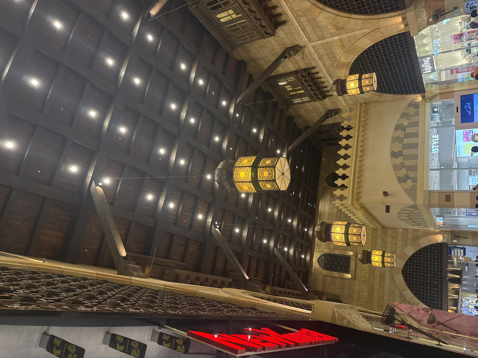 The image captures the interior of a building in Dubai, United Arab Emirates, with an architectural style reminiscent of traditional Middle Eastern design. The scene is dominated by a dark wooden coffered ceiling with numerous recessed lights. Large, ornate hanging lanterns with intricate yellow and green patterned glass are suspended from the ceiling by dark ropes. The walls are constructed of light-colored stone blocks, with decorative arched windows and wooden balconies featuring carved details. A large red illuminated sign is visible on the left side of the image, partially obscuring the stone wall. Below the balconies, storefronts with glass facades are visible, including signs for "iSTYLE" and "Kipling," indicating a commercial or shopping environment. A few people are faintly visible in the lower part of the image, near the storefronts, suggesting a public space. The overall impression is one of opulent and culturally inspired interior architecture.
