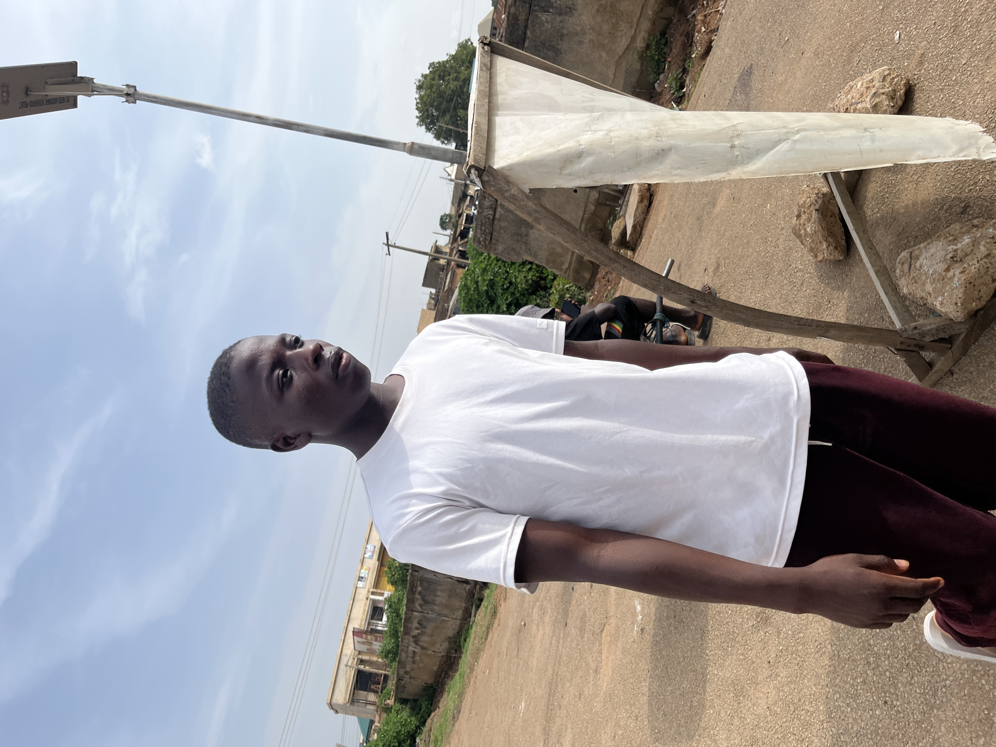A young Black male stands facing right, looking away from the camera, in an outdoor, urban setting. He is wearing a white t-shirt and maroon pants. His left foot is raised slightly. He is standing on a paved surface, possibly a road, which appears to be uneven and somewhat worn. In the background, there are buildings with a mix of concrete and corrugated metal roofing, and some vegetation. A tall, slender light pole with a solar panel fixture is visible to the right of the frame. In the foreground, a makeshift wooden structure holds a white, somewhat tattered fabric. Several rocks are placed around the base of this structure. The sky is partly cloudy and light blue. The overall impression is of a casual outdoor scene in a developing urban area. The location is identified as Rafin Par, Nigeria.