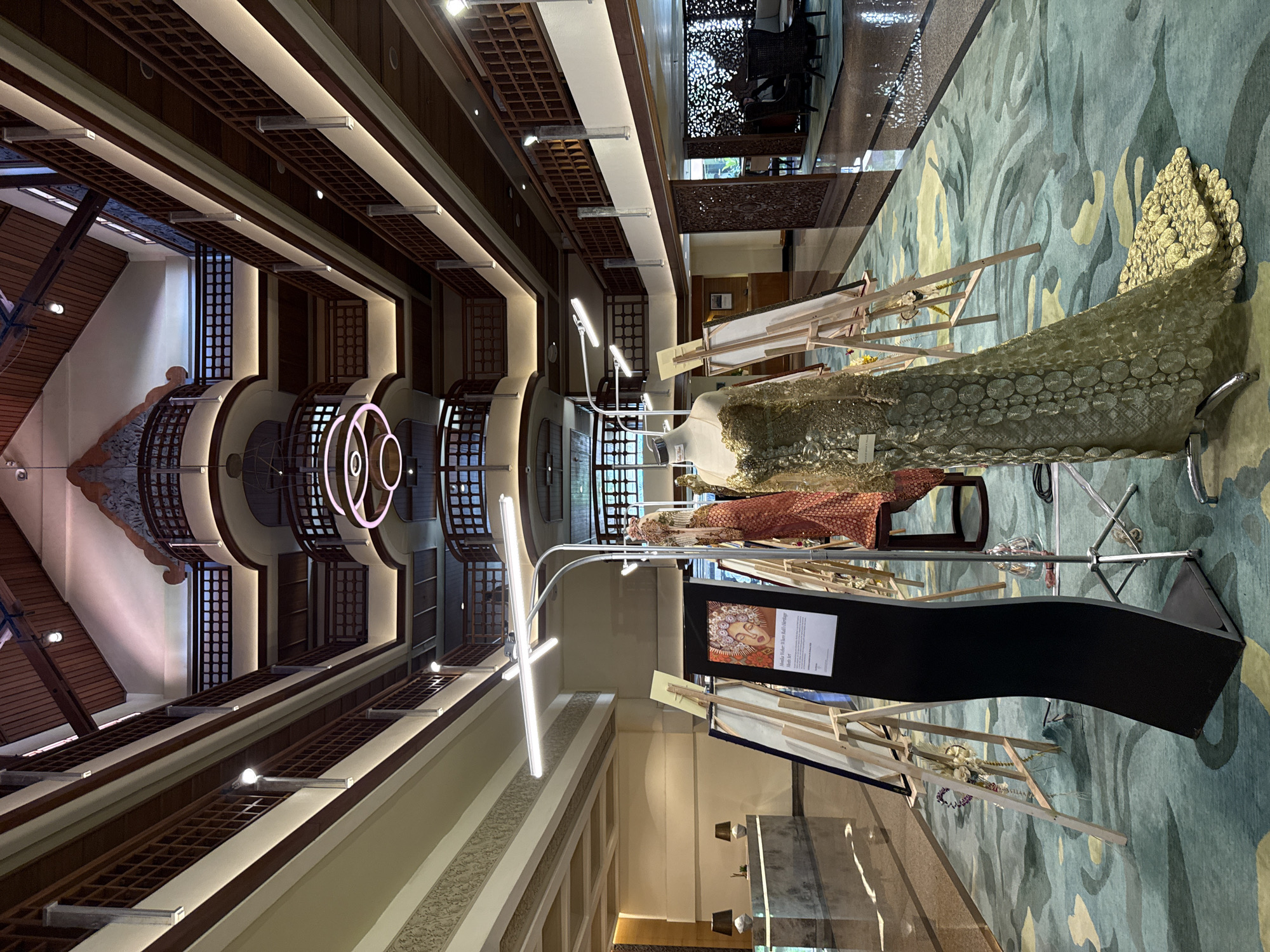 The image depicts the interior of a multi-story building in Nusa Dua, Indonesia, likely a hotel or resort lobby. The architecture features dark wooden latticework and light-colored walls with recessed lighting. A distinctive circular neon art installation is visible in the central atrium.
Several mannequins displaying ornate, possibly traditional, clothing are positioned on a patterned blue and green carpet. Easels supporting artwork and informational displays are situated nearby. A black display panel is also present. The scene appears to be part of an art exhibit or cultural presentation within the building.