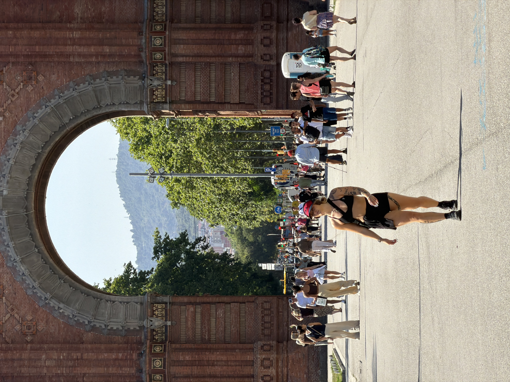 The image captures a sunlit scene at the Arc de Triomf in Barcelona, Spain. A large brick triumphal arch dominates the background, framing a view of a tree-lined avenue with distant hills under a clear blue sky. In the foreground, a woman with tattoos and a black bikini top and shorts walks towards the camera. She is wearing a red baseball cap and black boots. Other pedestrians are visible further down the avenue, walking in both directions. Some are dressed casually in summer attire, while others carry bags or wear backpacks. Traffic lights and street signs are present along the side of the road. The ground is paved asphalt with some faint chalk markings visible in the lower portion of the image. The overall atmosphere appears to be a busy, pleasant day in a public urban space.