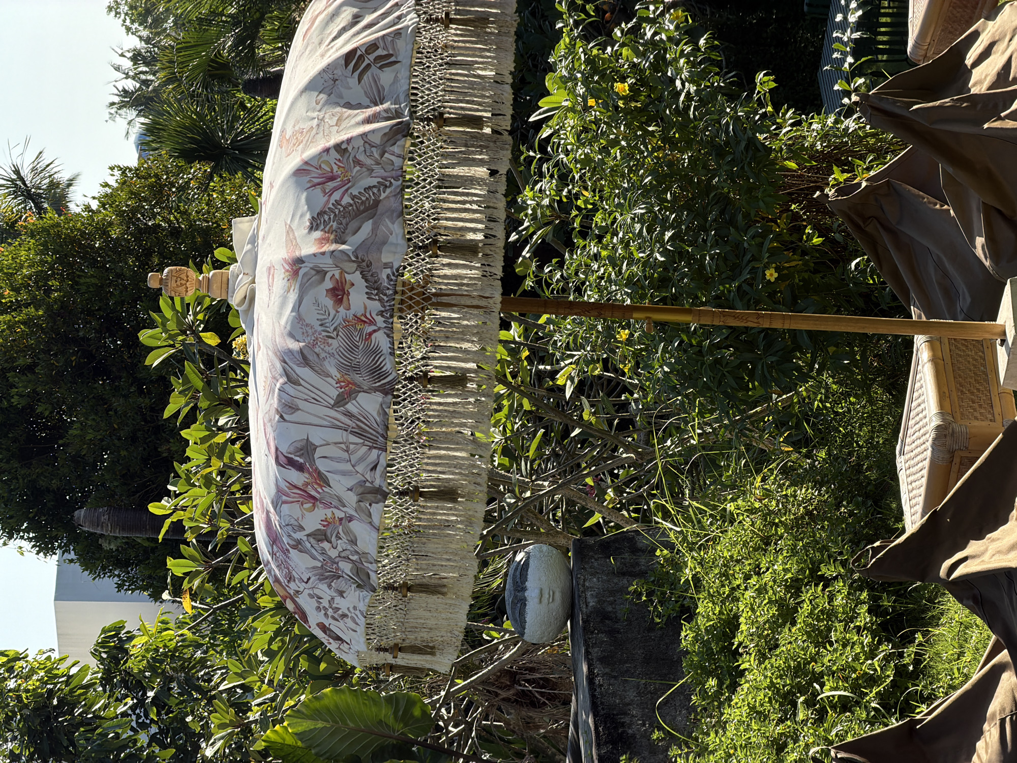 A brightly lit outdoor scene in Mengwi, Indonesia, features a large, decorative parasol as the central subject. The parasol canopy displays a tropical floral pattern with white ground, accented by reddish-pink, green, and brown foliage and flowers. Its lower edge is adorned with a double layer of fringed tassels, incorporating a woven lattice pattern above the longer tassels, and small, dark decorative elements. A carved wooden finial crowns the parasol's peak, supported by a light brown wooden pole that exhibits some carved details.
The surrounding environment is lush with various green tropical vegetation, including large trees with dense foliage in the background and smaller shrubs and bushes in the midground and foreground. A single yellow flower is visible amongst the green leaves in the midground. In the upper left background, a portion of a modern, light-colored building with a sharp corner is visible through the trees. A bare, textured tree trunk (possibly a palm) stands vertically in the upper center-right background. The sky, visible at the top, is clear and light blue.
To the left of the parasol pole, a grey and white decorative sculpture resembling a stylized face or head rests on a weathered concrete ledge or wall. In the immediate foreground, two dark brown, fabric-covered beanbag-style chairs are partially visible on the ground, accompanied by a small, light-colored rectangular table or stool made of wicker or rattan. The scene is illuminated by strong, natural sunlight, casting distinct shadows and highlighting the textures of the various elements. No people are present in the image.