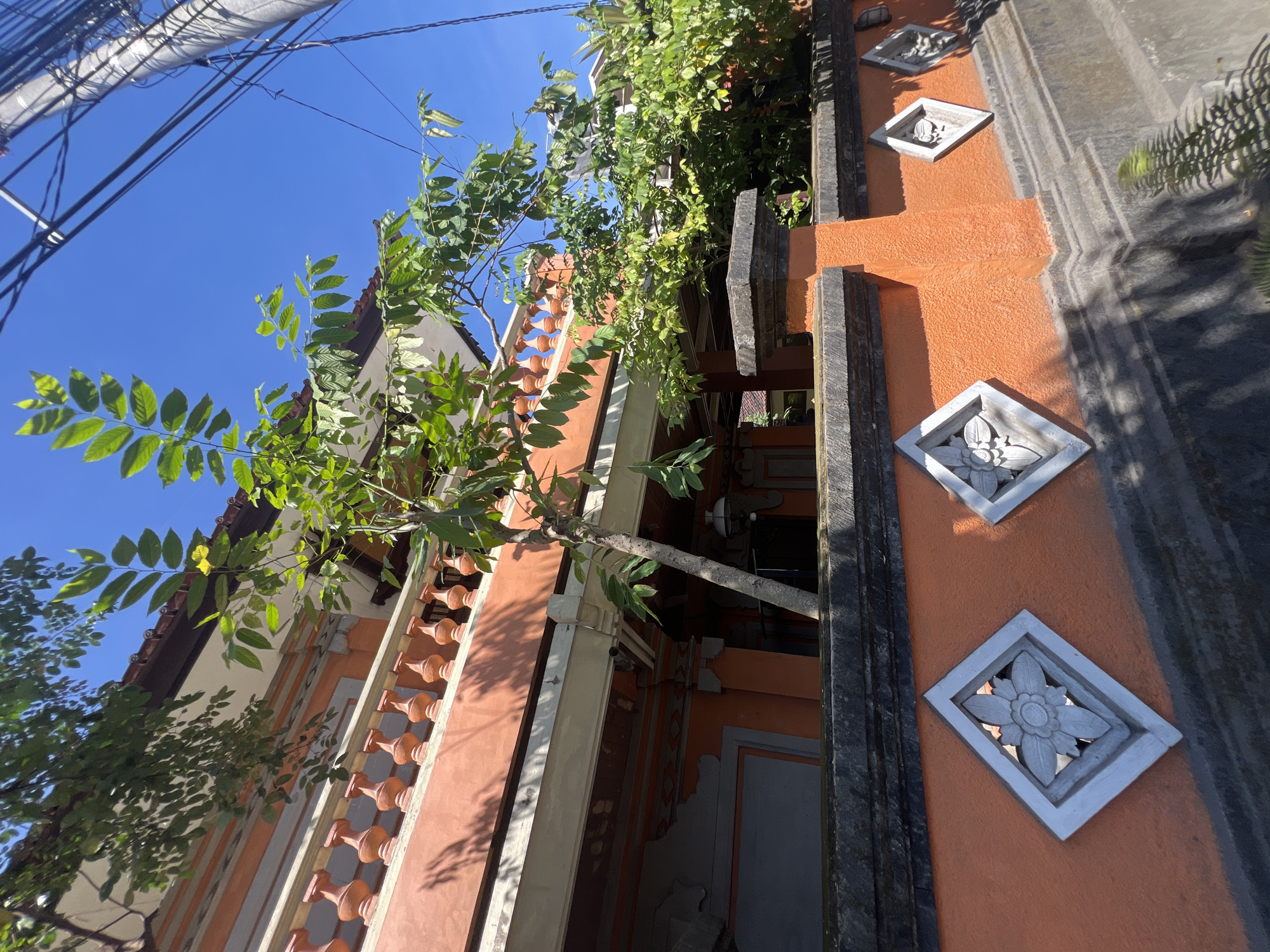 This is a medium-shot, low-angle, exterior, eye-level photograph of a bright orange, Balinese-style building and wall in Sukawati, Indonesia. The building has a light-colored second story with a red-tiled roof and a decorative terracotta balustrade. A small tree with green leaves grows in front of the building, reaching up towards the balcony. The wall in the foreground is also bright orange and features several diamond-shaped decorative reliefs with floral patterns. A dark gray stone cap runs along the top of the wall. Overhead, a utility pole and thick electrical wires are visible against a clear blue sky. The scene is illuminated by direct sunlight, casting shadows on the wall and ground. There are no people or animals visible in the frame.