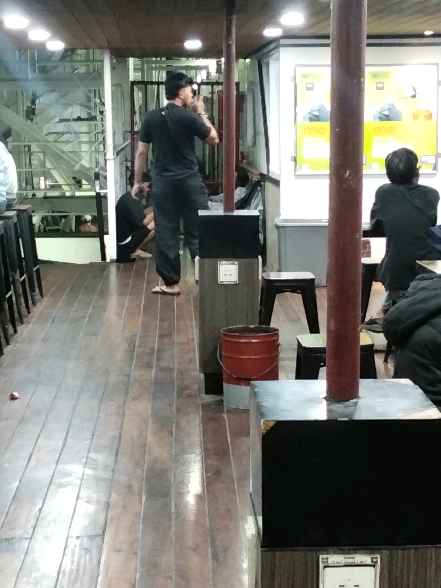The image depicts the interior of a ferry cabin, possibly on a route within Batam, Indonesia. The cabin features a wooden floor and ceiling, with fluorescent lighting. Several individuals are visible. In the foreground, a man in a black t-shirt and dark pants stands facing away from the camera, holding a phone to his ear. To his left, another person is seated on the floor, also in dark clothing. Further back, a person wearing a dark jacket is seated facing away from the camera, possibly at a table with another individual. In the background, posters with yellow and green elements are displayed on a white wall. The cabin contains seating areas with dark stools and tables. A red metal bucket and black cylindrical structures are also present. The overall impression is that of a functional, albeit somewhat dated, ferry interior.