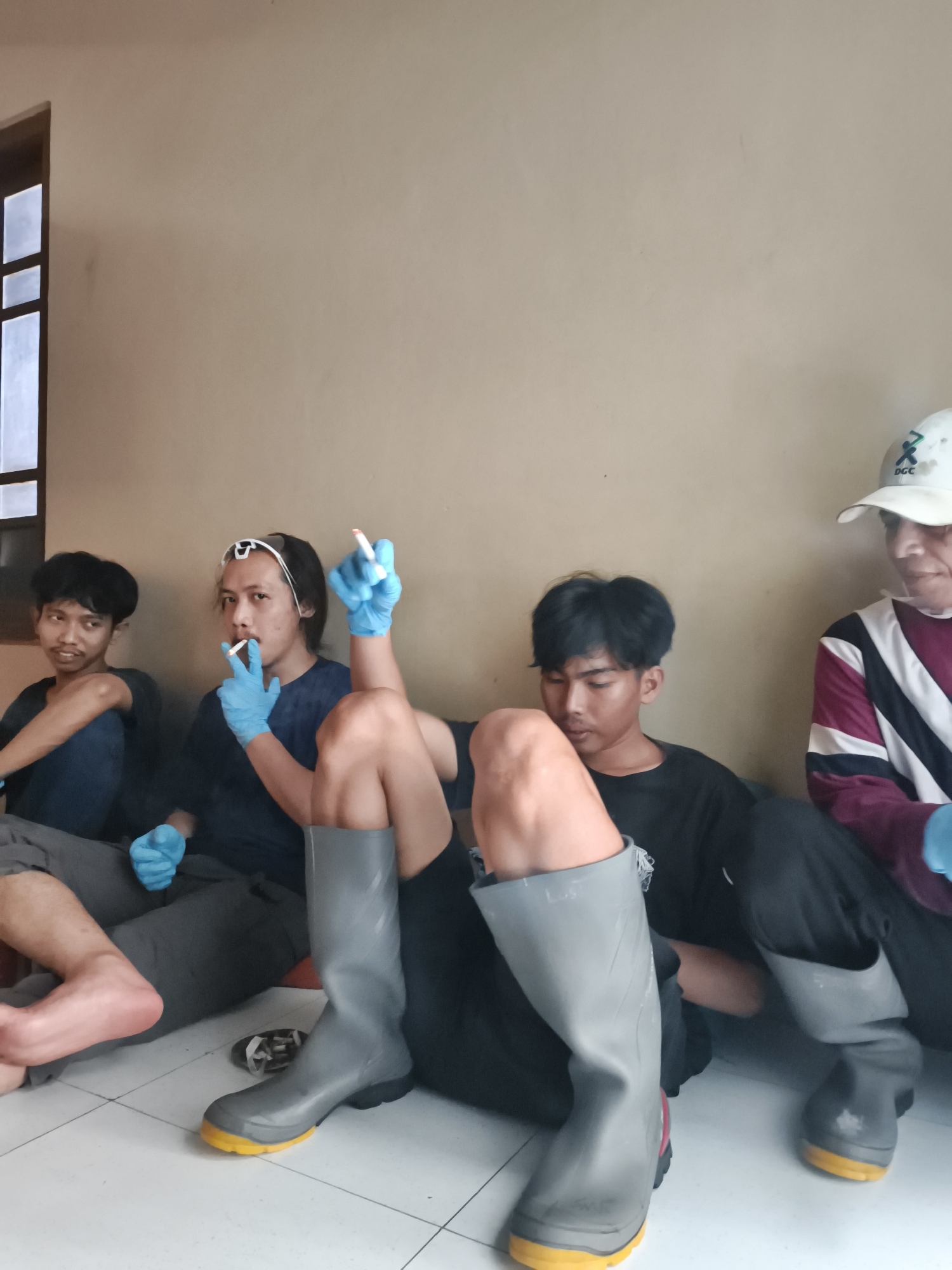 In Pekalongan, Indonesia, four individuals are seated on the floor against a plain, beige wall. Two individuals in the foreground are wearing large, grey rubber boots with yellow soles. One of these individuals is wearing a black t-shirt and is looking downwards. The other individual is also wearing dark clothing. The two individuals in the background appear to be younger men. The man on the far left is wearing a dark blue shirt and looking to the side. The man next to him, in the center of the image, is wearing a dark blue t-shirt and is smoking a cigarette. He has blue gloves on both hands and a clear plastic band across his forehead. The man on the far right is wearing a white baseball cap with a logo and a burgundy and white striped shirt, and he also appears to be wearing blue gloves. A window with multiple panes is visible on the left side of the image. The floor is tiled with a white and grey grid pattern.