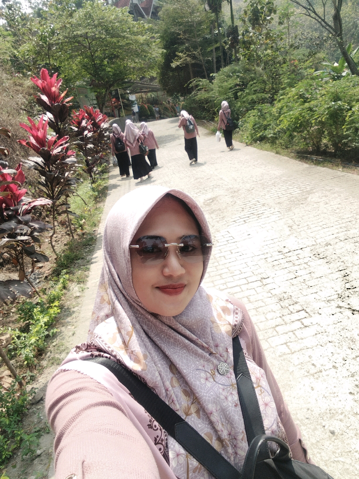 In Kaliwedi, Indonesia, a woman in a pink hijab and sunglasses takes a selfie on a paved path. She is wearing a light pink sweater and a patterned hijab. A black bag strap is visible over her shoulder. Behind her, several other women in similar attire are walking away from the camera down the path. Red-leafed plants line the left side of the path. The environment is outdoors with trees and foliage.