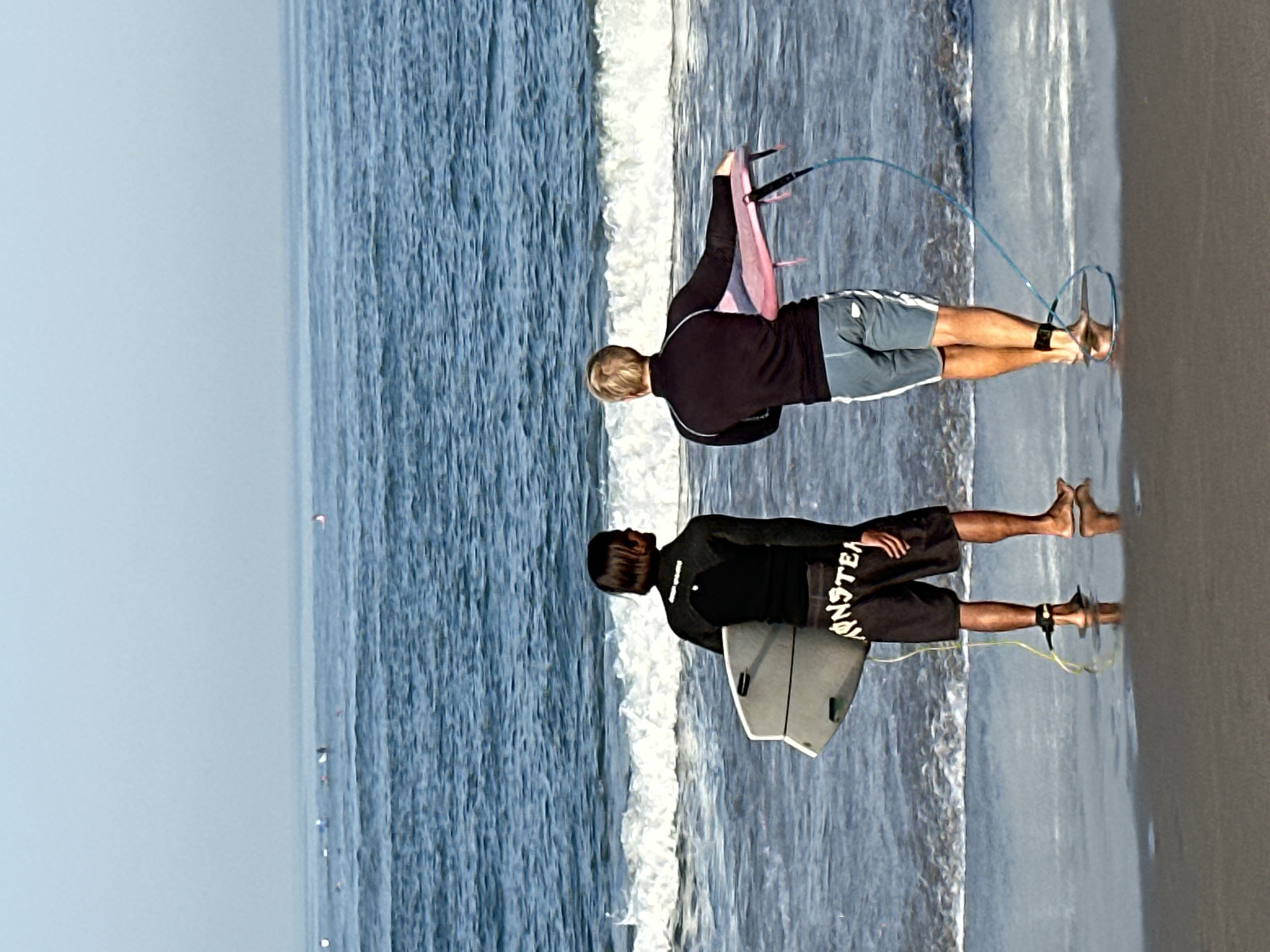 Two individuals, viewed from behind, stand at the edge of the ocean on a sandy beach. The person on the left, wearing a black wetsuit and black shorts with white lettering, holds a light-colored surfboard. The person on the right wears a black long-sleeved shirt and blue-gray shorts and holds a pink board with straps. Both individuals have a leash attached to their ankles, connecting them to their respective boards. The ocean is blue with moderate waves breaking near the shore. The sky is a hazy light blue, indicating a potentially overcast or distant horizon. The sandy beach is wet from the receding tide, reflecting the sky and the figures. In the distance, several other surfers can be seen in the water. The scene depicts a typical day for surfing activities.