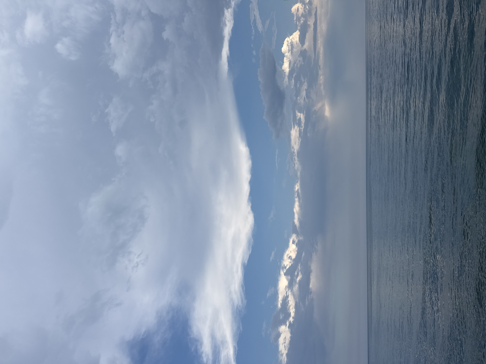 The image depicts a wide expanse of the Black Sea meeting a cloudy sky. The water is calm, with subtle ripples reflecting the muted light from above. The sky is filled with a variety of clouds, ranging from wispy cirrus formations high up to heavier, darker clouds lower down. Patches of blue sky are visible between the clouds, and some rays of sunlight are breaking through in the distance. The horizon line is clearly defined where the sea meets the sky. There are no people, structures, or specific landmarks visible, suggesting an open, natural environment. The scene is likely located in or near Partenit, Ukraine, given the context of the Black Sea.
