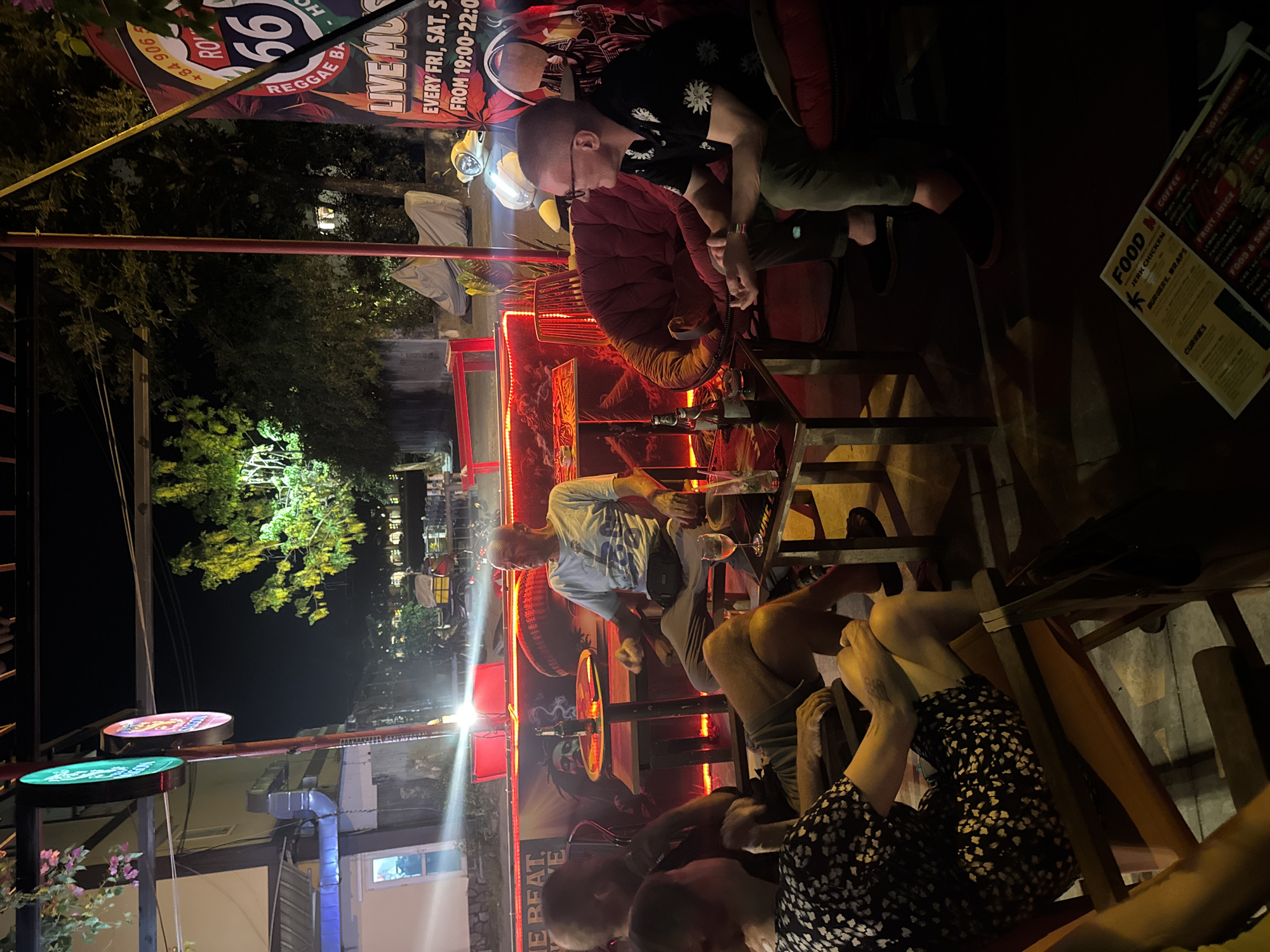 The image shows an outdoor scene at night at a bar or restaurant, likely in Đà Nẵng, Vietnam. Several people are seated at tables. A sign reading "Route 66 Reggae Bar" is visible. A food menu lists items such as Jerk Chicken, burgers, wraps, curries, coffee, and fruit juice. The bar has red lighting and some trees are in the background. There are multiple chairs and tables visible. One person is wearing a black shirt with white floral designs. Another person is wearing a red shirt.