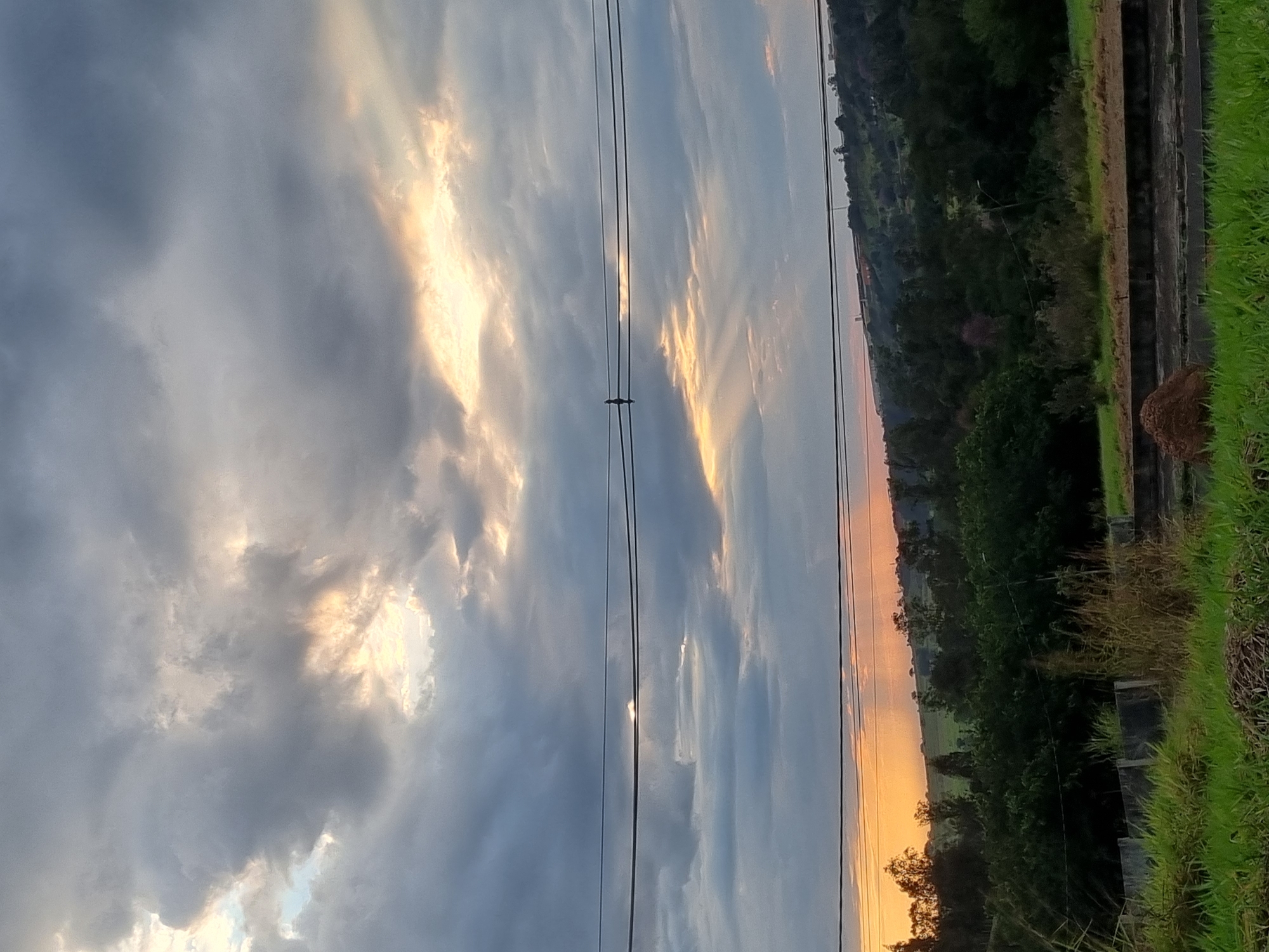 The image shows a cloudy sky with rays of sunlight breaking through, casting a dramatic effect over a landscape. In the foreground, there is lush green grass and some low concrete structures. In the middle ground, a dense line of trees and foliage stretches across the horizon, with a few taller trees interspersed. Above the trees, power lines are visible, with a small bird perched on one of them. The background features rolling hills with more trees and some distant buildings, suggesting a semi-rural or suburban environment. The time of day appears to be either sunrise or sunset due to the warm light filtering through the clouds. The image does not provide specific landmarks or details to pinpoint the exact location within Laranjal Paulista, Brazil.
