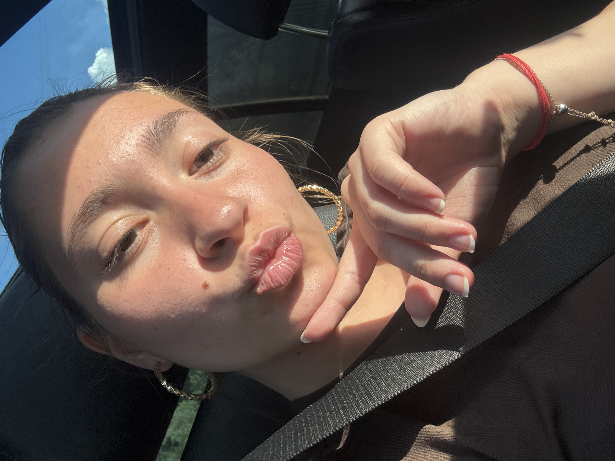The image is a close-up, indoor, selfie-style photograph of a young woman with her hand on her chin, puckering her lips. She is wearing large hoop earrings and a red string bracelet on her wrist. A seatbelt is visible across her chest. The background suggests she is in a car, with a dark interior and a glimpse of a blue sky with clouds through the sunroof. The lighting indicates it is daytime. There is no indication of the location being the city of Dolno Varshilo, Bulgaria.