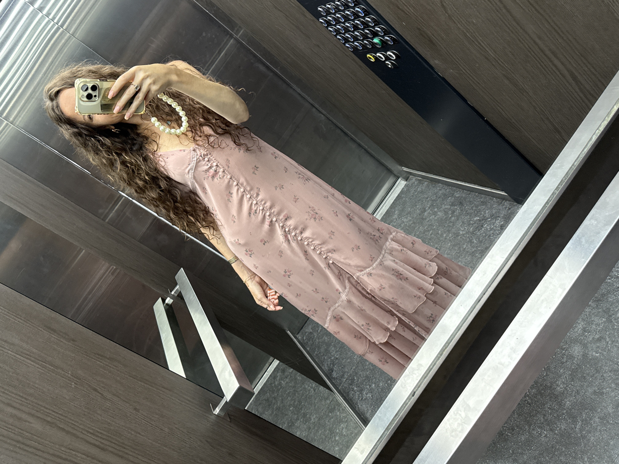 A woman with long, curly brown hair is taking a selfie in an elevator. She is wearing a long, pale pink floral dress with ruffles and buttons down the front. She is also wearing a pearl necklace. The elevator has brushed metal walls and a dark wood accent panel with buttons. The floor is grey and textured. The reflection in the elevator doors and walls show the woman and the elevator interior. The scene appears to be in an urban environment, possibly in the city of Khimki, Russia, given the context.