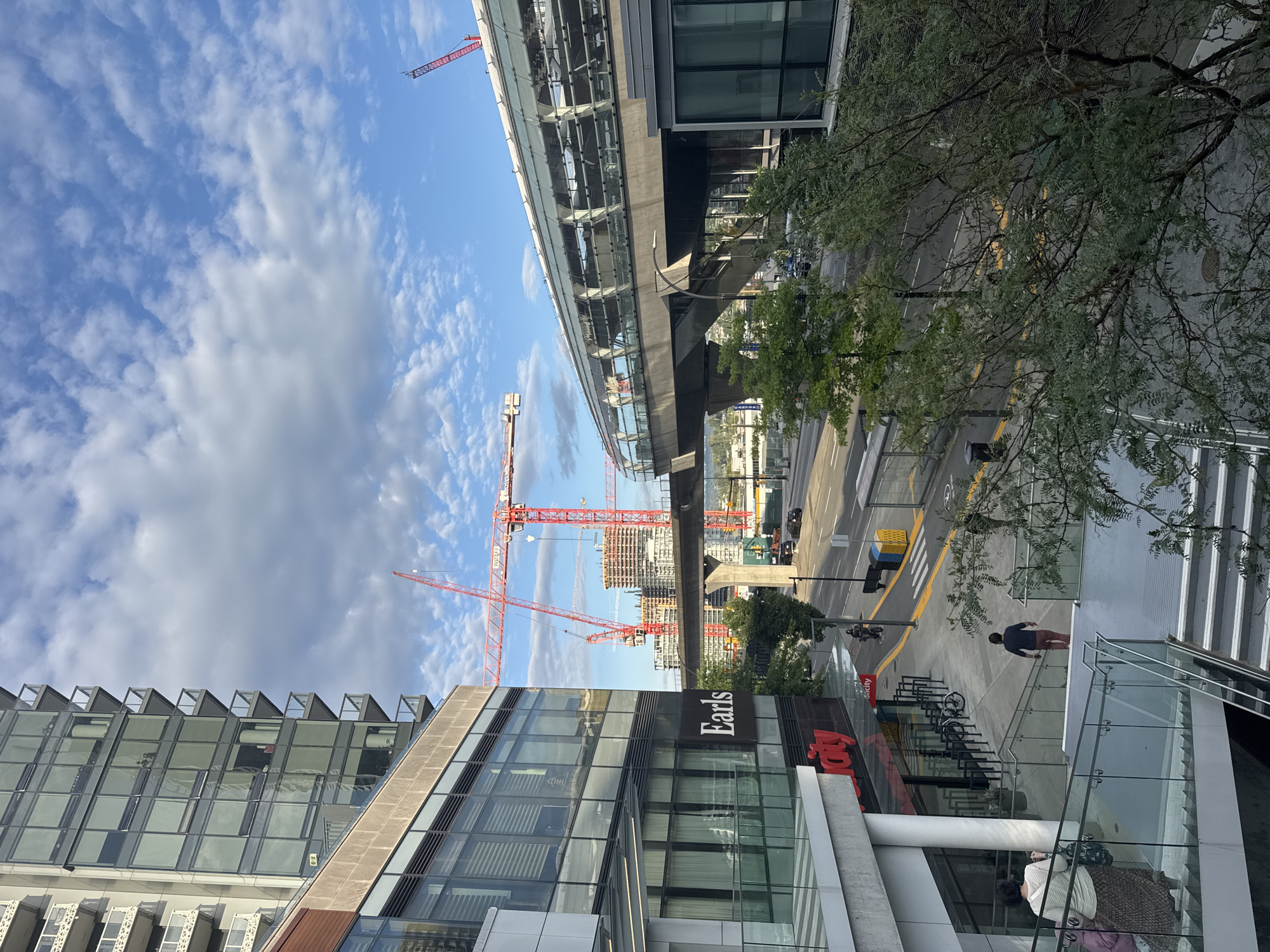 The image depicts an urban street scene in Burnaby, Canada, with a focus on ongoing construction projects and public transit infrastructure. In the foreground, a modern building with large glass windows and a sign reading "Earls" is visible. Below this, a red sign with white lettering that appears to say "city" is partially obscured. A woman is walking down a set of stairs on the left, and a man in a dark shirt and reddish pants is walking up the stairs on the right.
In the mid-ground, a SkyTrain guideway, characterized by its curved glass enclosure and concrete supports, spans across the scene. Beyond the SkyTrain, multiple construction cranes, predominantly red, are prominently featured, indicating significant development. One tall, dark-colored building under construction is visible behind the SkyTrain. A street runs parallel to the SkyTrain, with several cars and a motorcycle visible. Traffic lights and street signs are present, and a pedestrian crossing with yellow markings is also depicted.
The background reveals a sky with scattered clouds and a bright blue expanse. Green trees are scattered throughout the scene, some appearing in planters near the buildings and others along the street. The overall impression is one of a bustling urban environment undergoing significant growth and modernization.