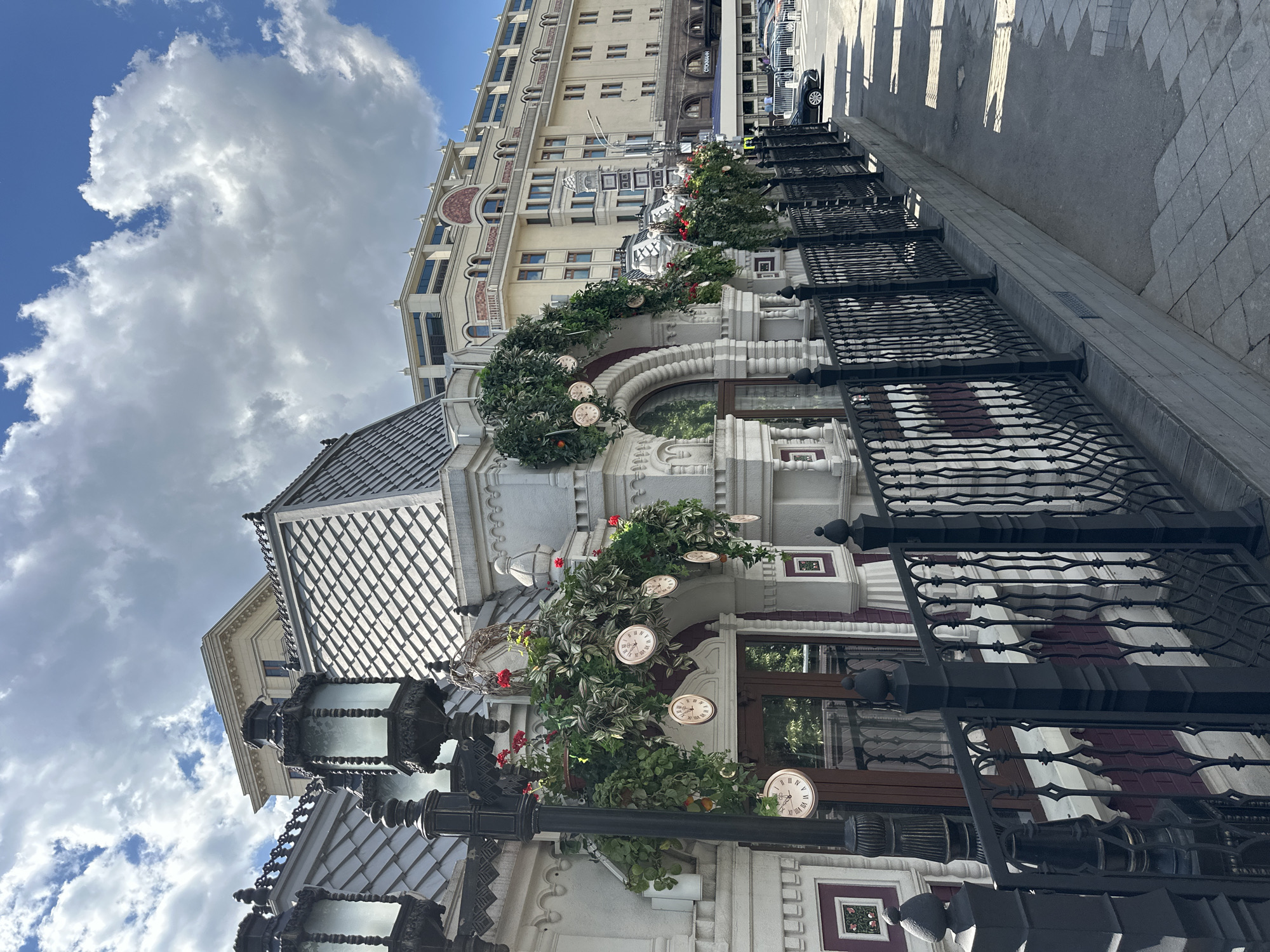 This image captures an elegant urban street scene in Moscow, Russia, taken from a low angle and looking upwards, providing a dynamic perspective. The setting features ornate architecture under a bright, partly cloudy sky.
In the foreground, a decorative building or storefront facade is prominent, distinguished by its light-colored stone or stucco and an intricate, possibly tiled, roof. This structure is heavily adorned with lush green foliage, interspersed with small red flowers and numerous decorative clock faces showing Roman numerals. These clocks are integrated into the greenery, creating a whimsical and charming display above arched windows. Ornate black street lamps with frosted glass are also visible, contributing to the classical aesthetic.
Behind this, a taller, multi-story building rises, featuring a light-colored facade with many windows and decorative elements, including circular accents. A partial sign on this building displays the text "CROMAN".
To the right, a black wrought iron fence with intricate patterns runs along a paved sidewalk, separating the buildings from the street. Beyond the sidewalk, a dark-colored car is visible on the street in the distance. Long, distinct shadows are cast across the sidewalk, indicating strong sunlight and suggesting it is daytime, likely morning or afternoon.
The sky is a vibrant blue with large, white, fluffy cumulus clouds, indicating fair weather. There is no specific activity or event taking place; the scene is a static view of a beautifully decorated streetscape. The overall impression is one of architectural beauty and sophisticated urban charm.