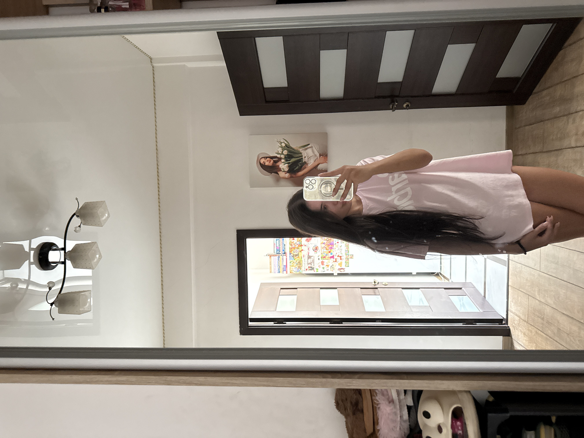 A young woman with long, dark hair takes a selfie in a full-length mirror. She is wearing a pale pink t-shirt dress and has a phone with a pop socket attached in her right hand. She is standing in what appears to be a bedroom or dressing area. To her left, a shelf unit is partially visible, holding various items including what looks like a plush toy with a cartoonish face. Behind her, a closed dark wood door with glass panes is visible, and to the left of that, another open wooden door leads to a hallway adorned with colorful posters. A framed picture of a woman in a hat holding tulips hangs on the wall to the right of the open door. The room has white walls and a wooden floor. The ceiling has a modern chandelier with three square glass shades.