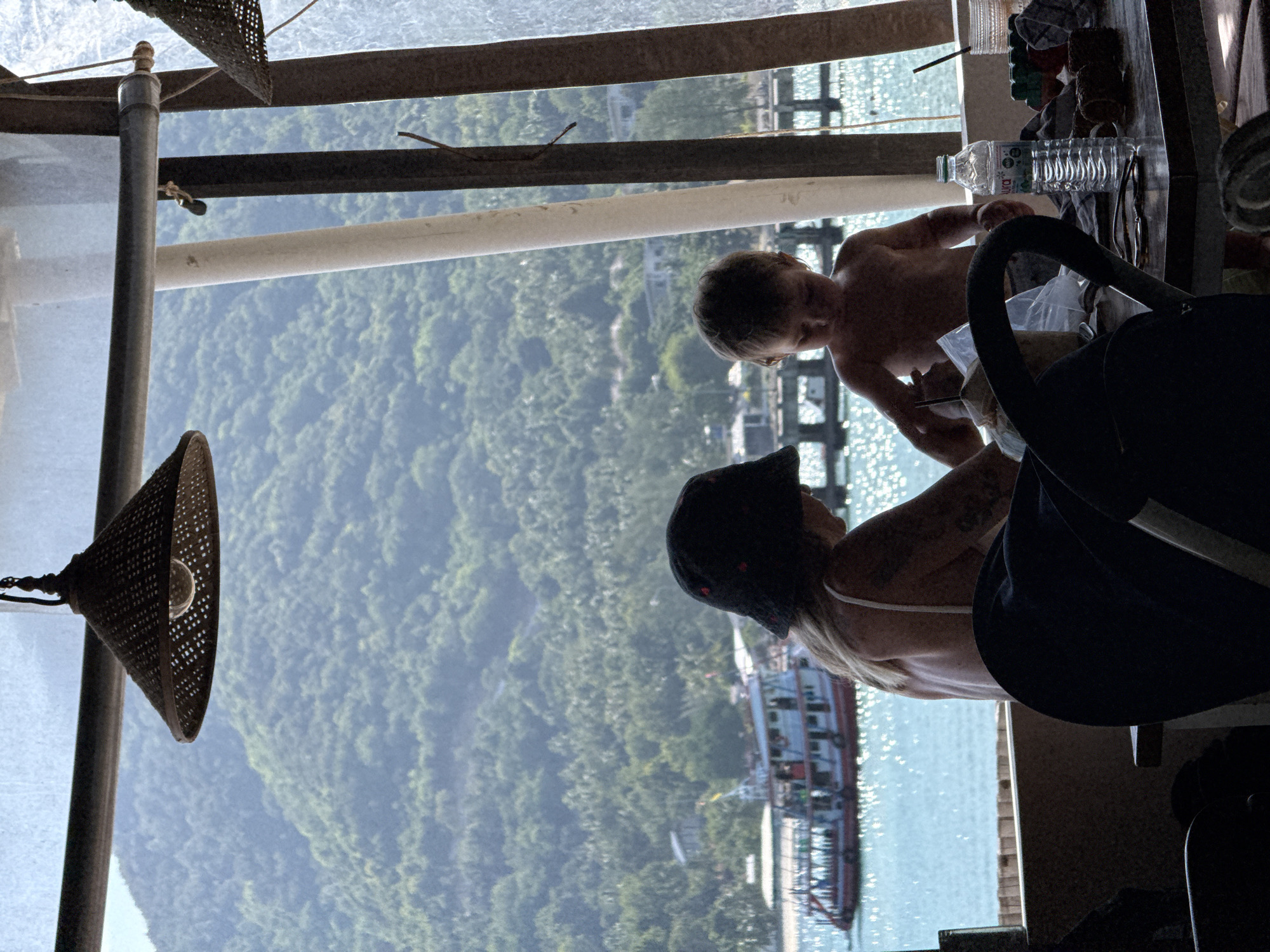 A family is enjoying a meal at a beachfront cafe on Ko Pha-ngan District, Thailand. A woman, wearing a black bucket hat, sits at a table with a young child. In the background, a boat sails across the turquoise waters of the Gulf of Thailand, with lush green mountains rising behind it. The cafe appears to be open-air, with woven light fixtures hanging from the ceiling. A bottle of Aura water and other items are on the table.