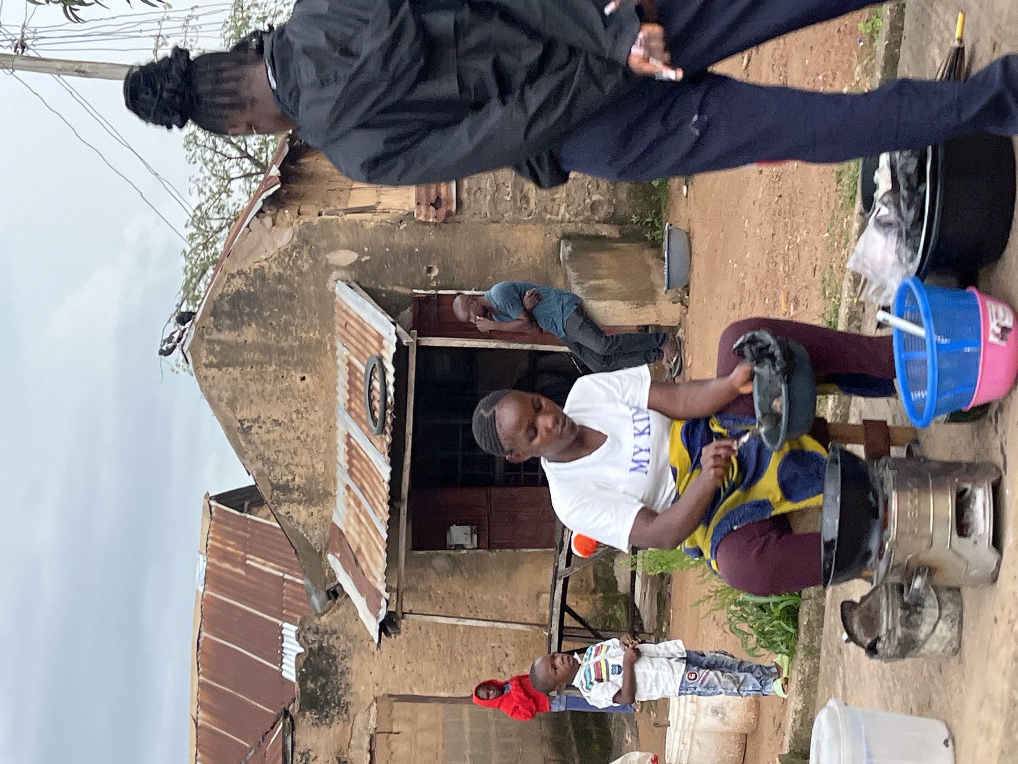 In Bukuru, Nigeria, an outdoor scene unfolds with a woman in a white t-shirt and a yellow and blue patterned wrap cooking over a portable stove. She is holding a dark bowl, seemingly preparing food. To her left, a young boy in a patterned shirt and blue jeans stands attentively. Behind them, another child in a red hooded sweatshirt is visible. A man in a teal shirt and dark pants is seated to the right of the cooking area, looking to the side. In the foreground, various containers and baskets are present, including a large black basin filled with plastic bags. The background features a building with a corrugated iron roof, typical of the region, and electrical wires overhead. The ground is composed of dirt and sand, indicating a rural or semi-urban setting within the city.