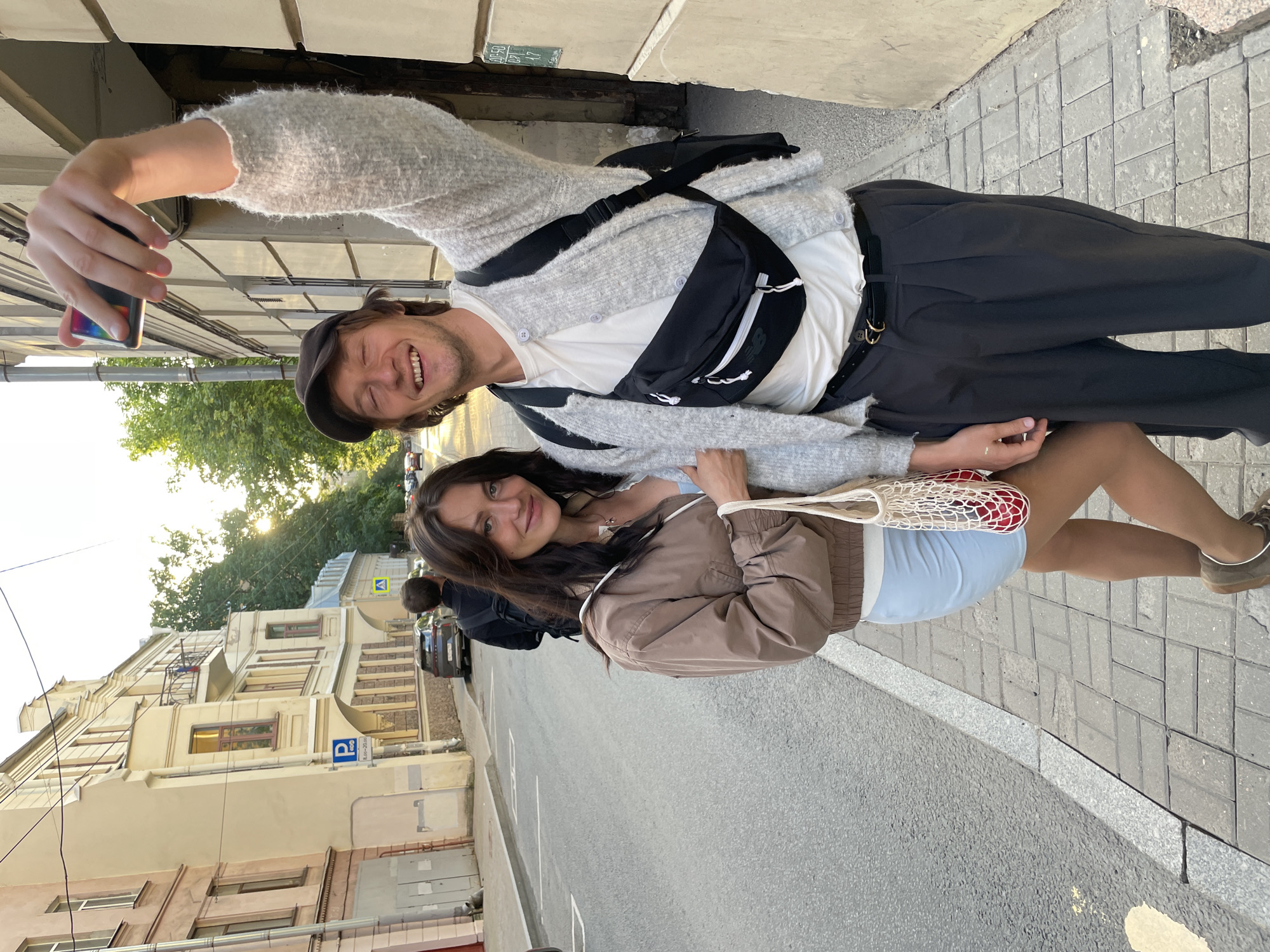 Two young adults, a man and a woman, are standing on a sidewalk on a street in Saint Petersburg, Russia, taking a selfie. The man, on the right, is wearing a light grey cardigan over a white t-shirt, dark grey pleated pants, a black belt, a black fanny pack, and a dark baseball cap. He is holding up a smartphone with his right hand, capturing the photo. The woman, on the left, is wearing a light brown bomber jacket over a light blue dress and is carrying a mesh bag containing red fruit. She is smiling towards the camera. In the background, there are buildings lining the street, a parked car, and trees. A street sign with a "P" symbol and Russian text is visible on a building. The sidewalk is paved with cobblestones. The sun is setting, casting a warm glow on the scene.